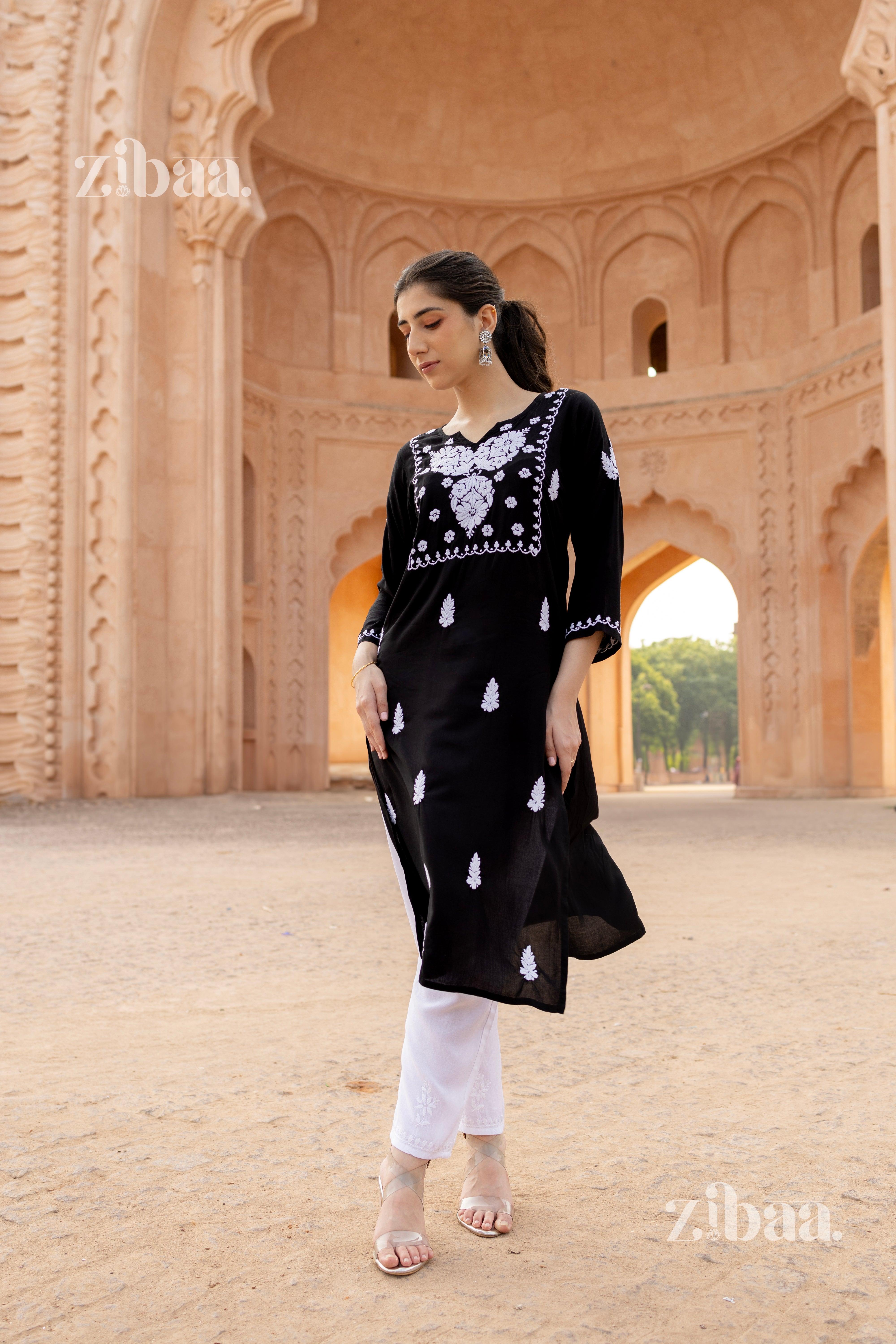 A woman wearing a black Chikankari kurti with white embroidery, styled with white pants, standing in front of an architectural backdrop.