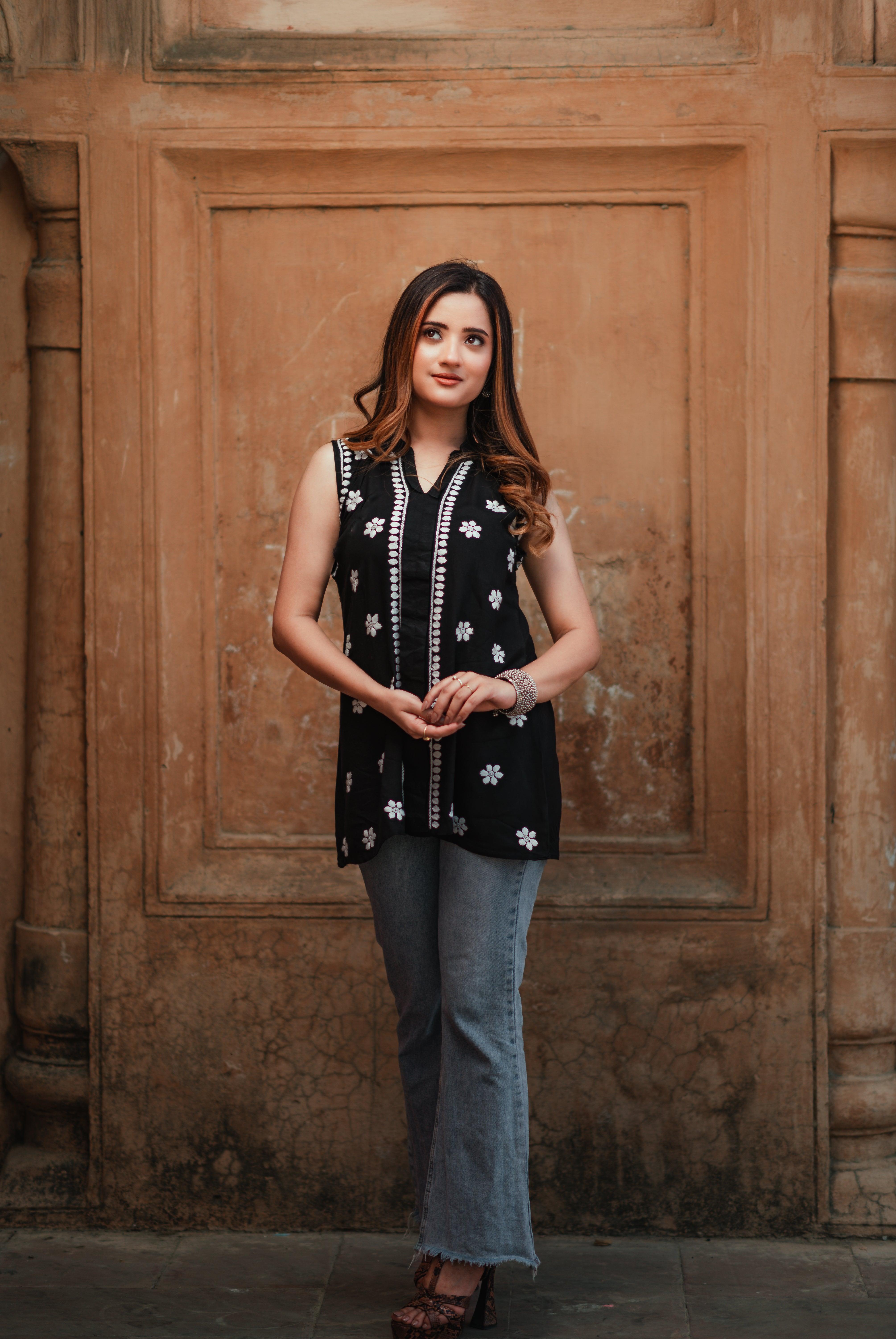 A woman in a sleeveless black Chikankari short kurti with white embroidery poses with hands gently clasped, in front of a rustic wall.