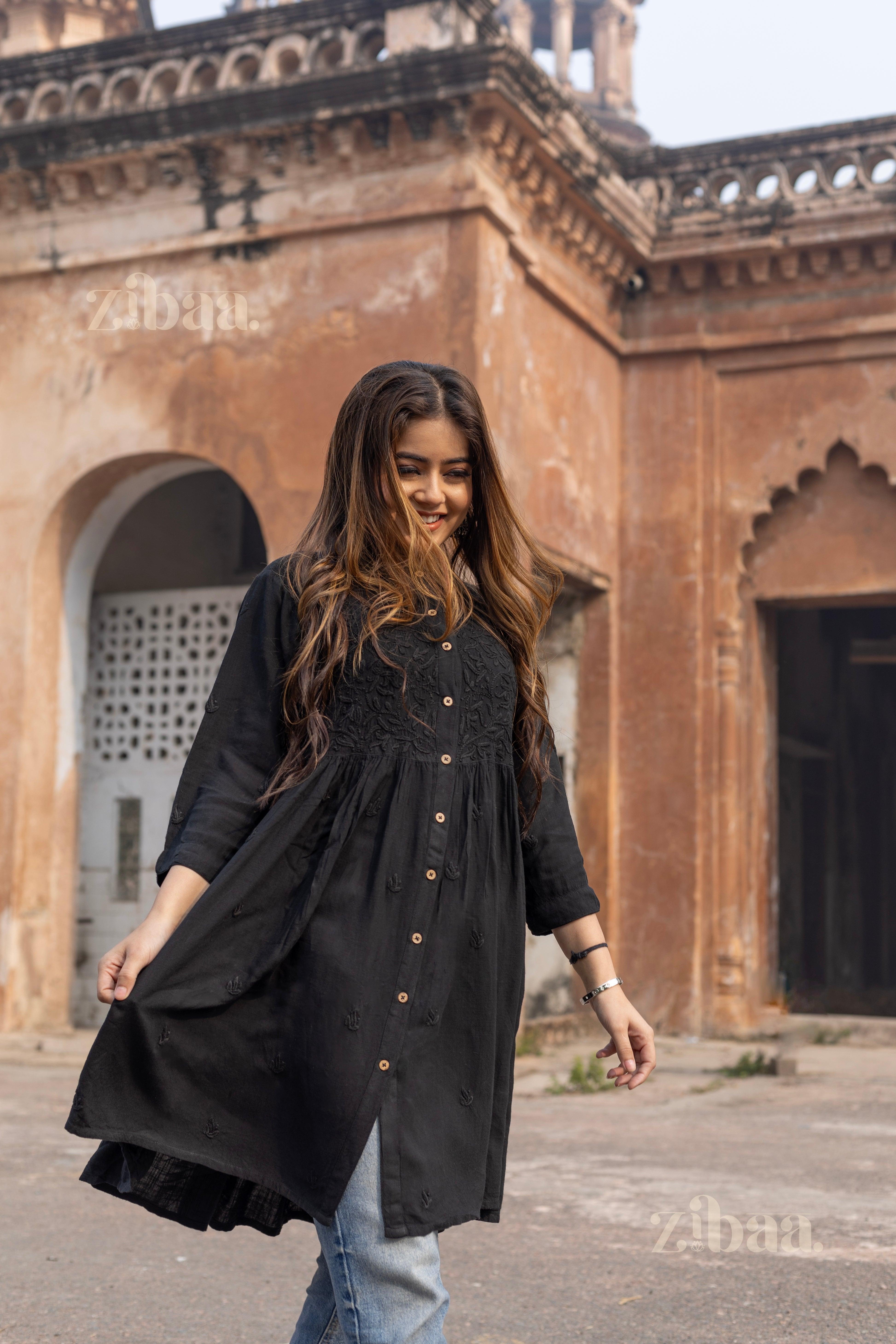 A woman joyfully twirls outdoors, wearing a black Chikankari top with intricate embroidery, paired with jeans and a smiling expression.