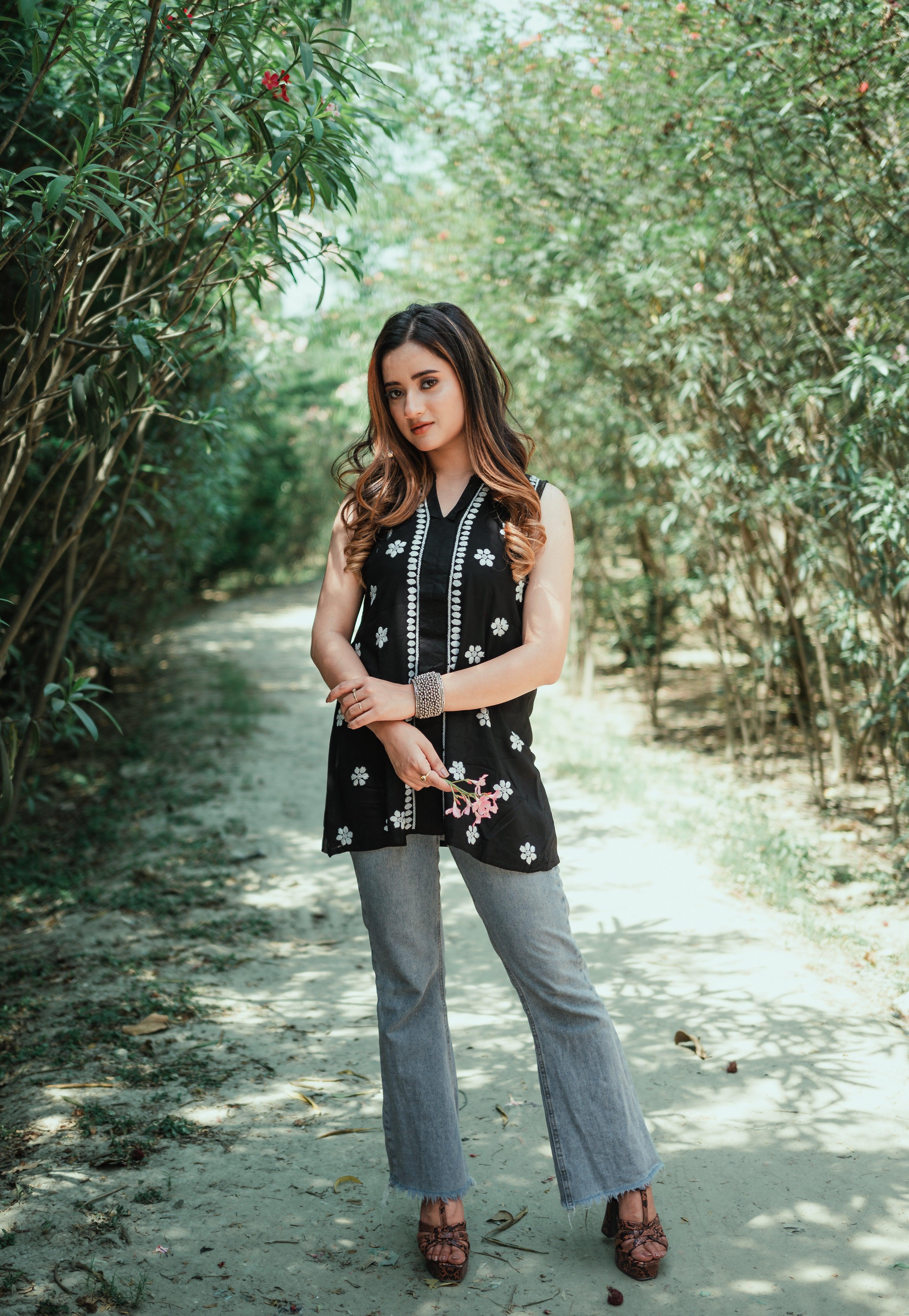 A woman wearing a sleeveless black Chikankari top with white embroidery stands on a leafy pathway, holding pink flowers in one hand.