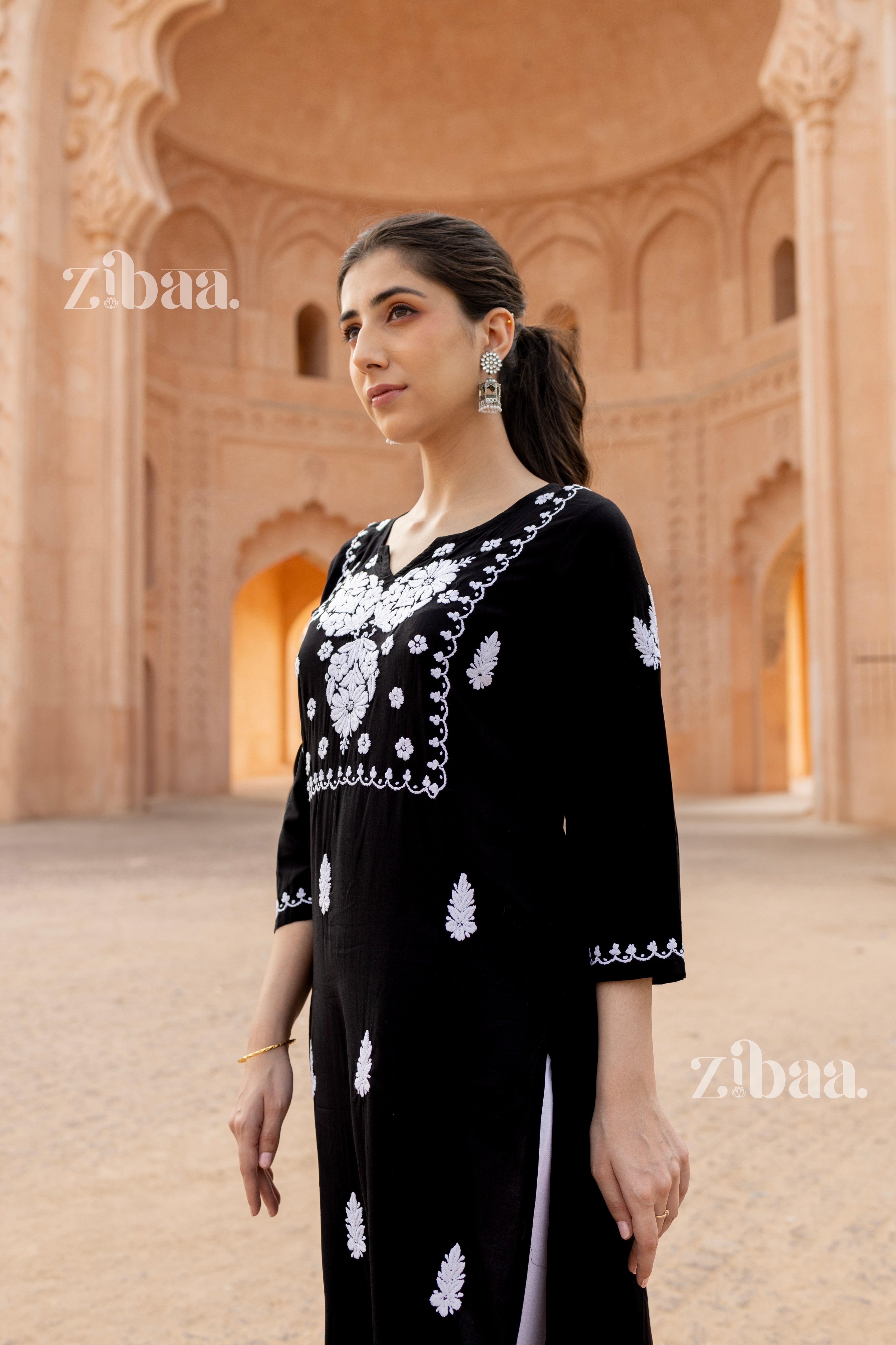 A woman wearing black and white kurti with lots of Chikankari embroidery stands holding herself against a heritage-inspired backdrop.