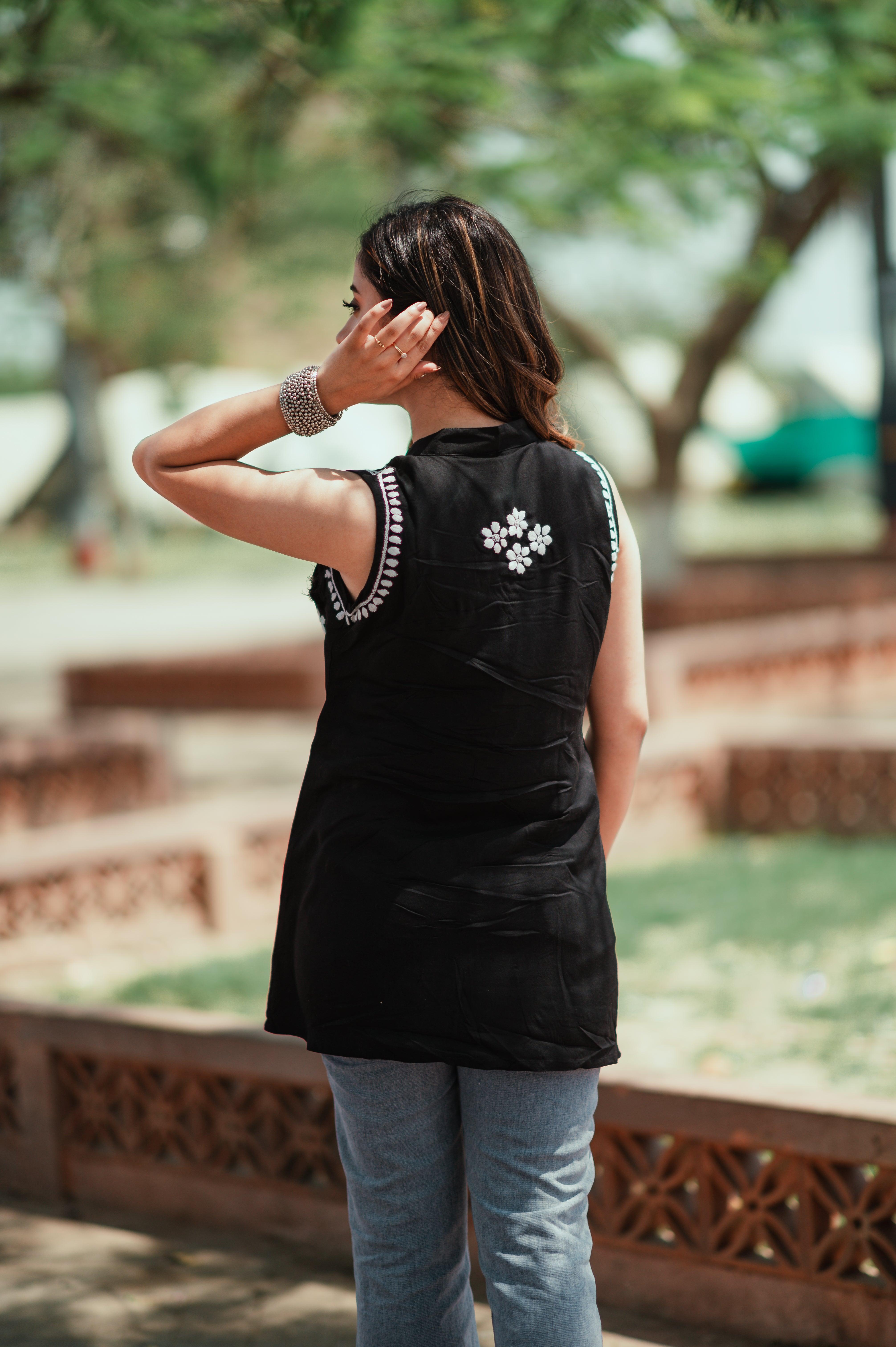 A woman wearing a black Chikankari short kurti with white embroidery stands outdoors, facing away, touching her hair with one hand