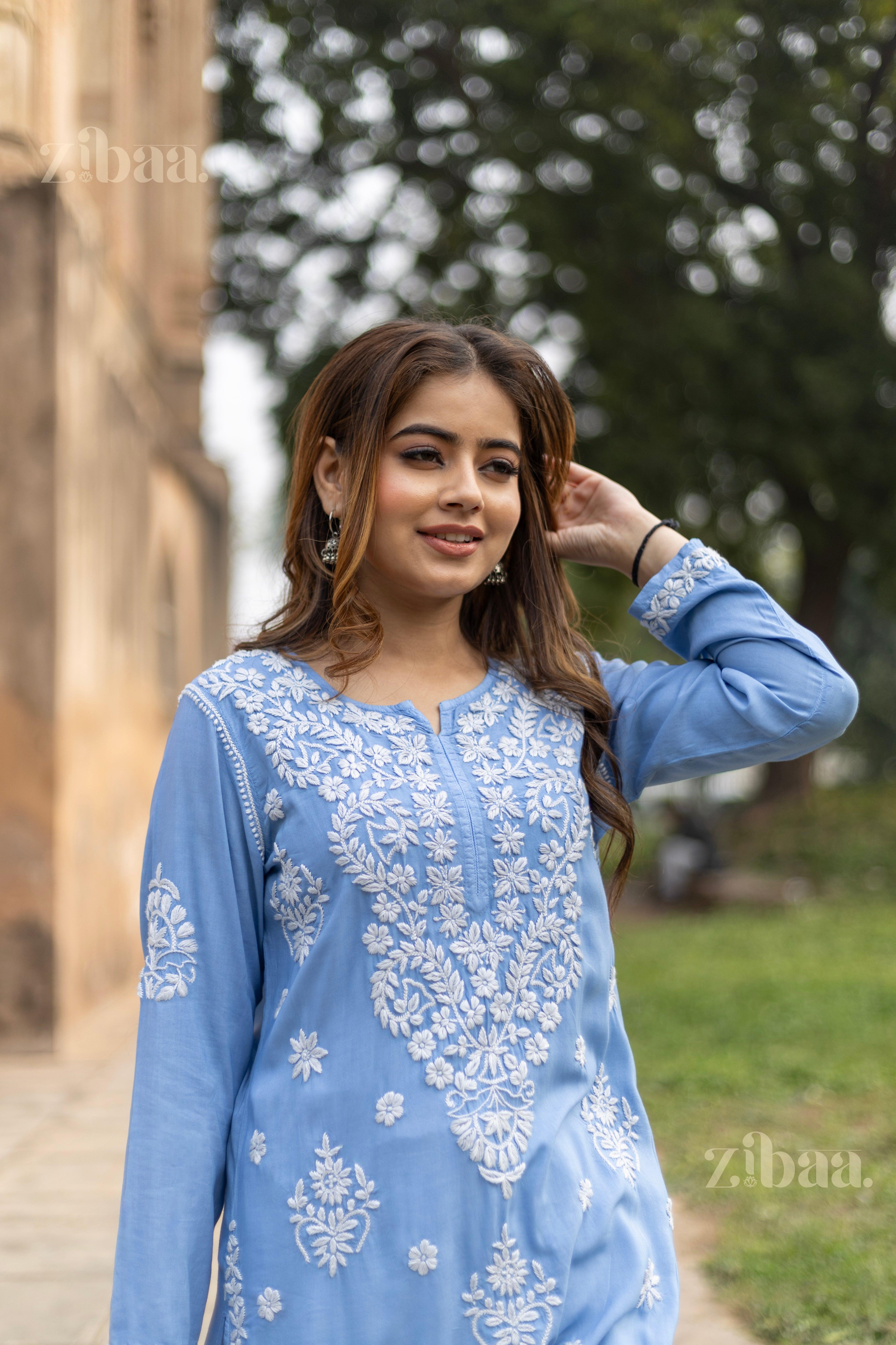 Model in the blue Lucknowi Kurti with delicate white embroidery poses outside gracefully against serene green and rustic backdrops.