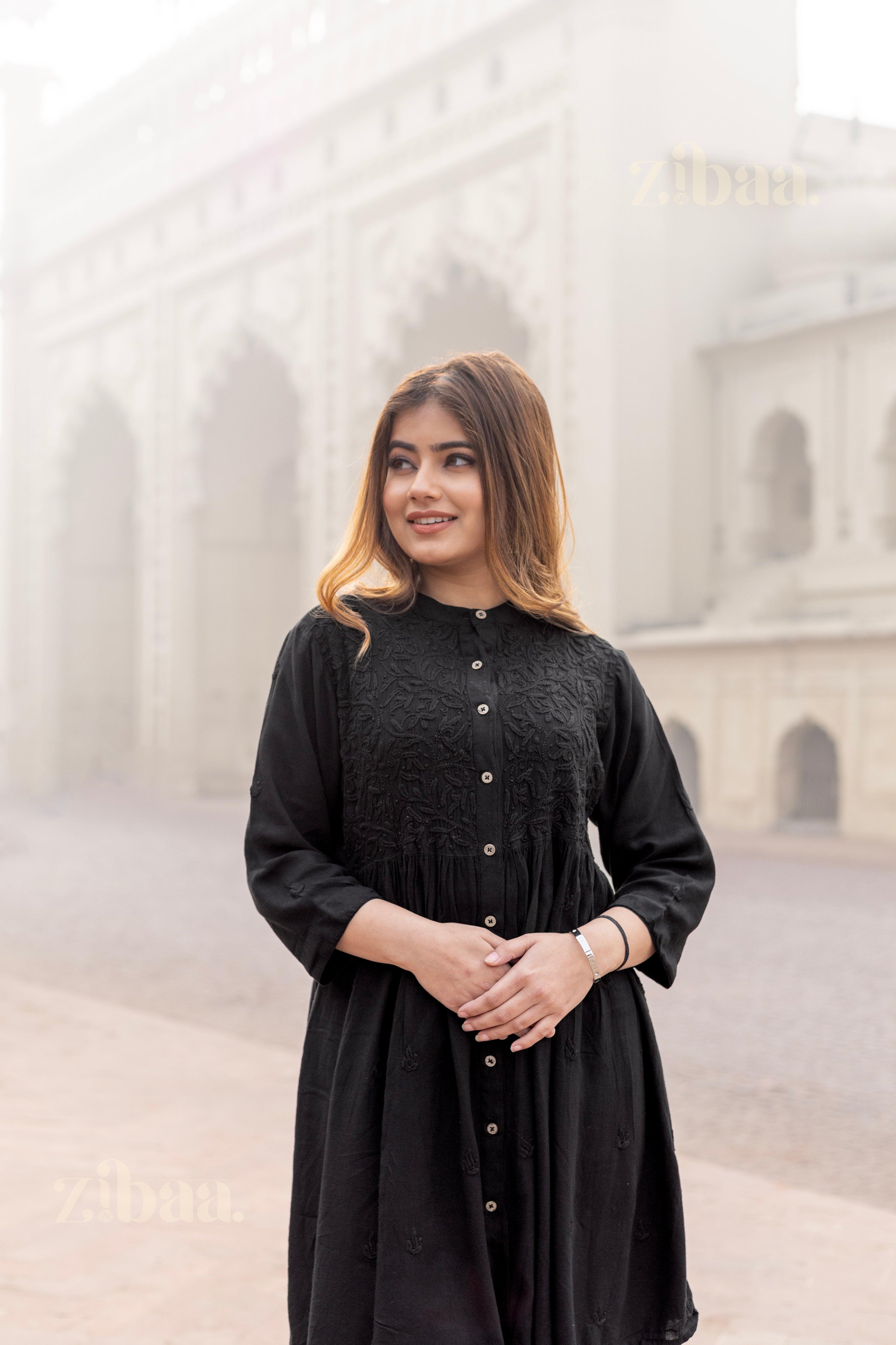 A woman stands outdoors in a misty backdrop, wearing a black rayon Chikankari Kurti for women, with her hands gently clasped.