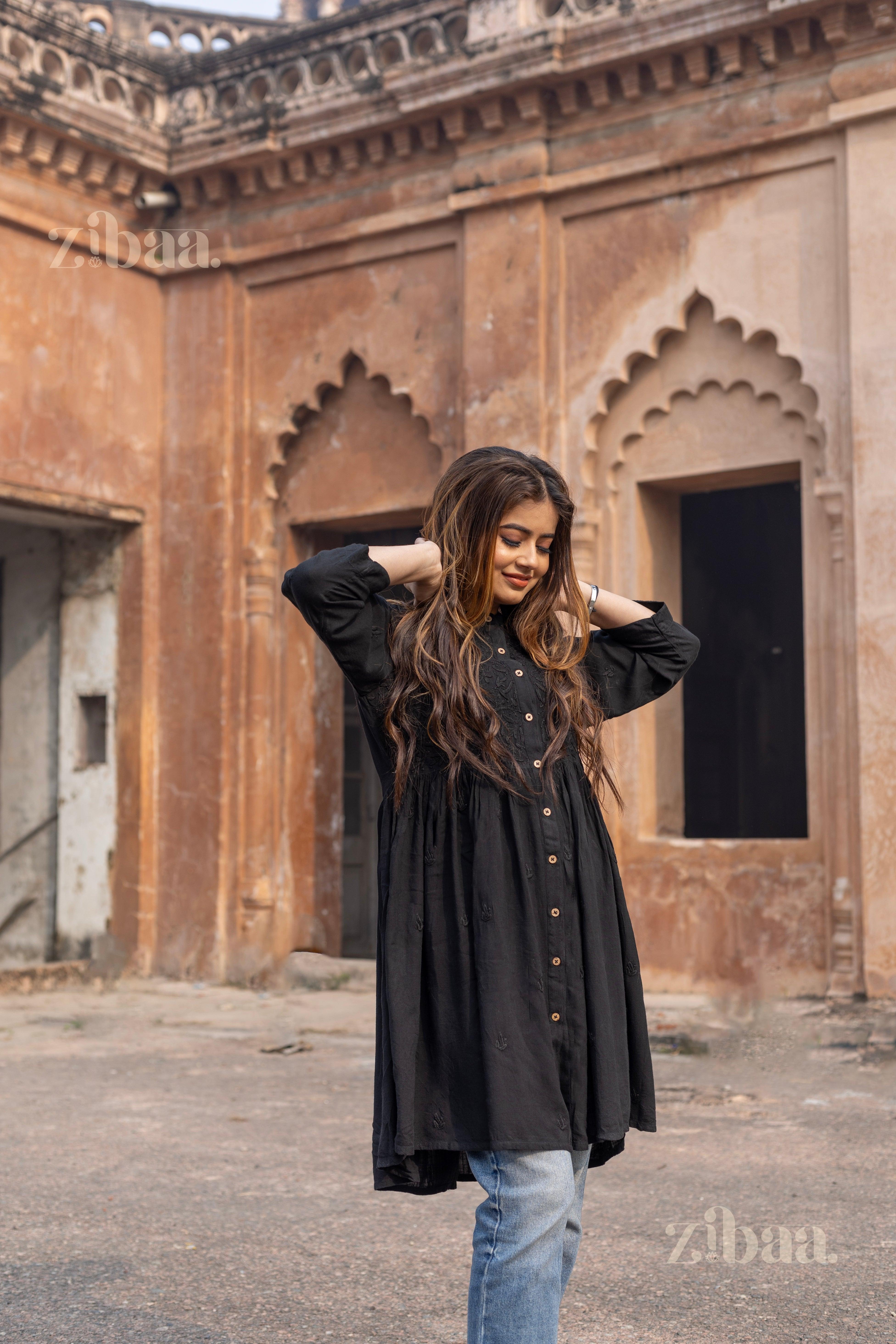 A woman adjusts her hair with a joyful smile, wearing a black Chikankari top with intricate embroidery, paired with jeans.