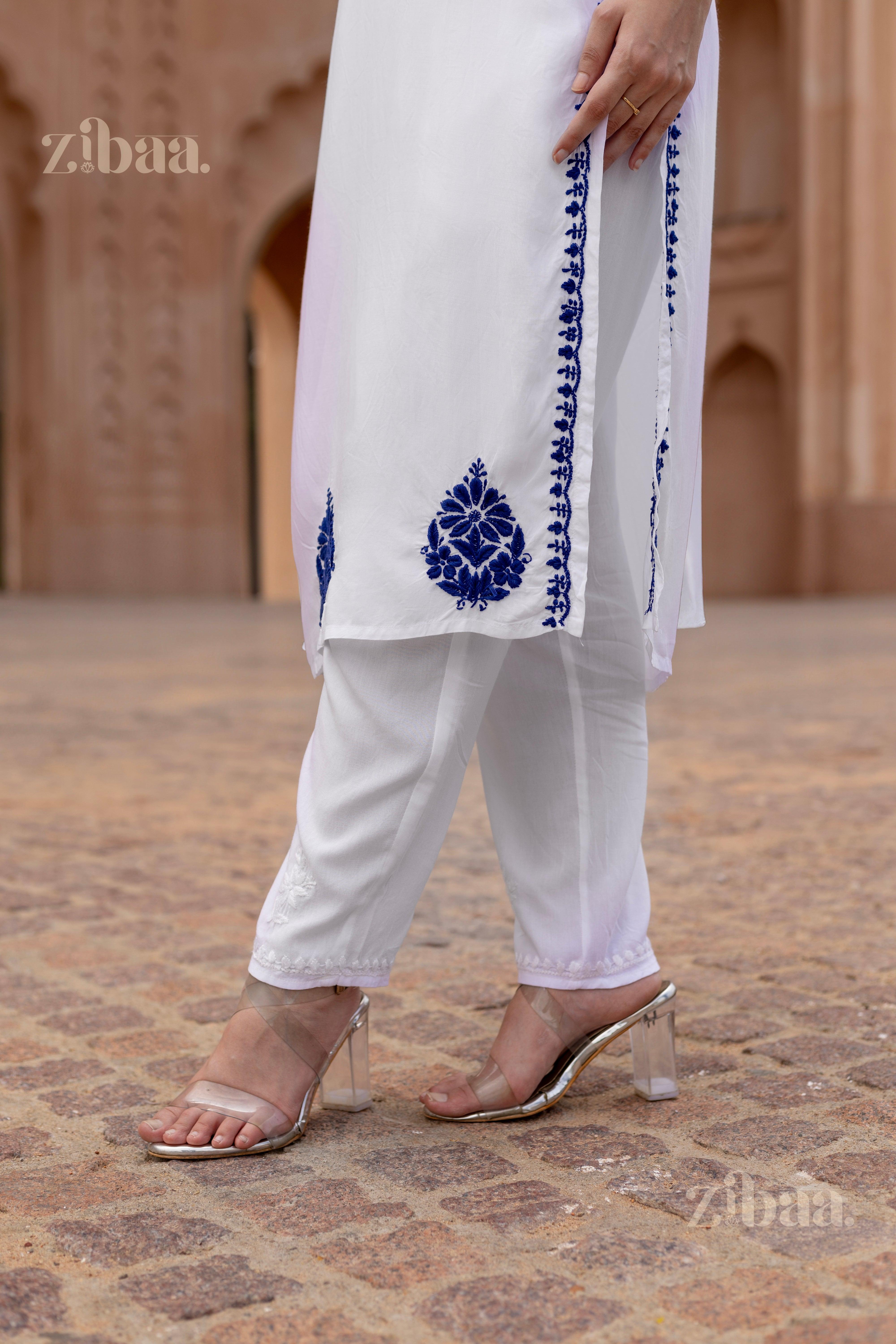 Close-up of a woman wearing a white Chikan Kurti with blue embroidery on the hem, paired with white pants and transparent heels, outdoors.