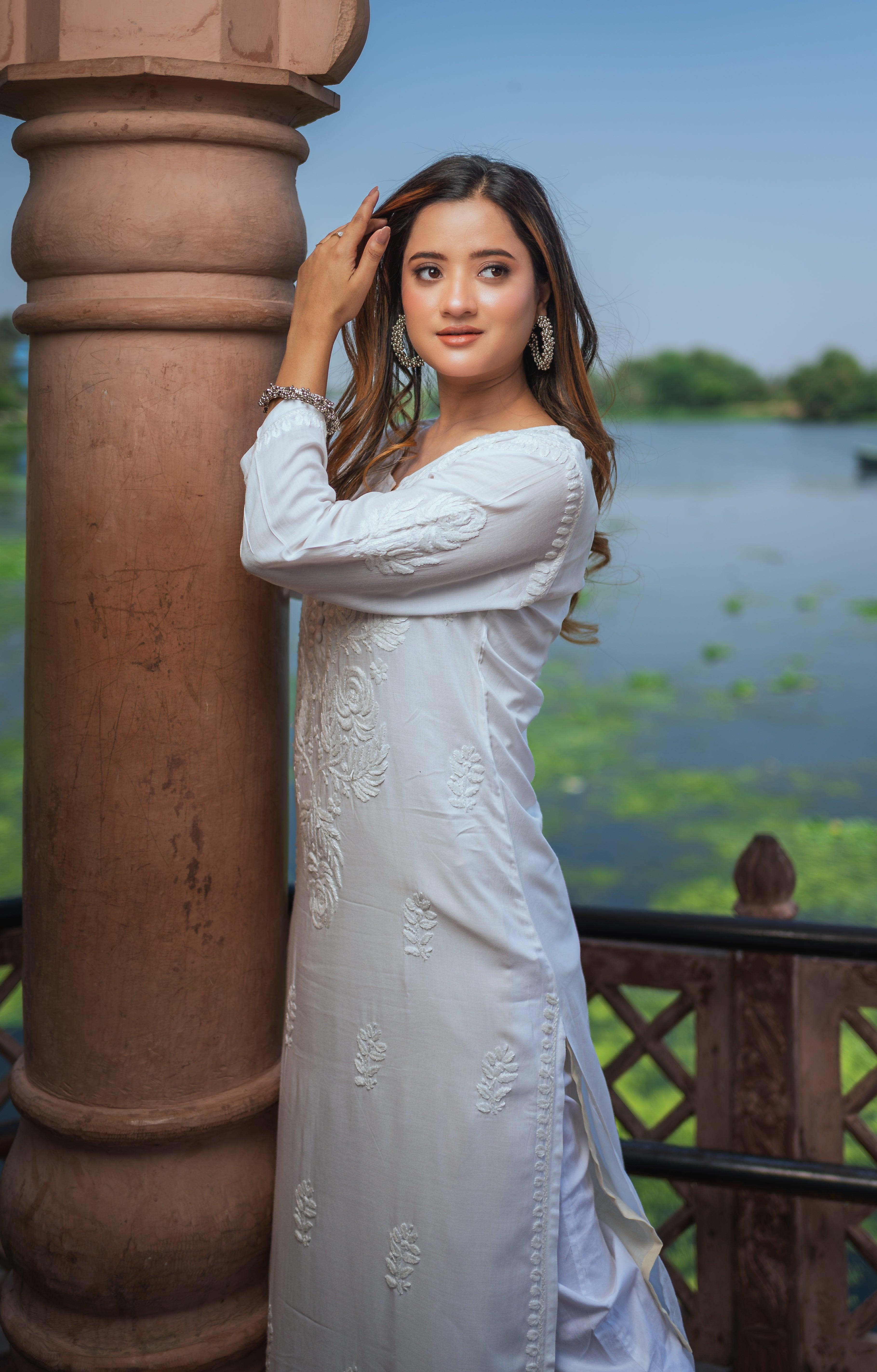 A woman in a white Chikankari kurti with delicate embroidery stands beside a pillar, touching her hair, and looking confidently.