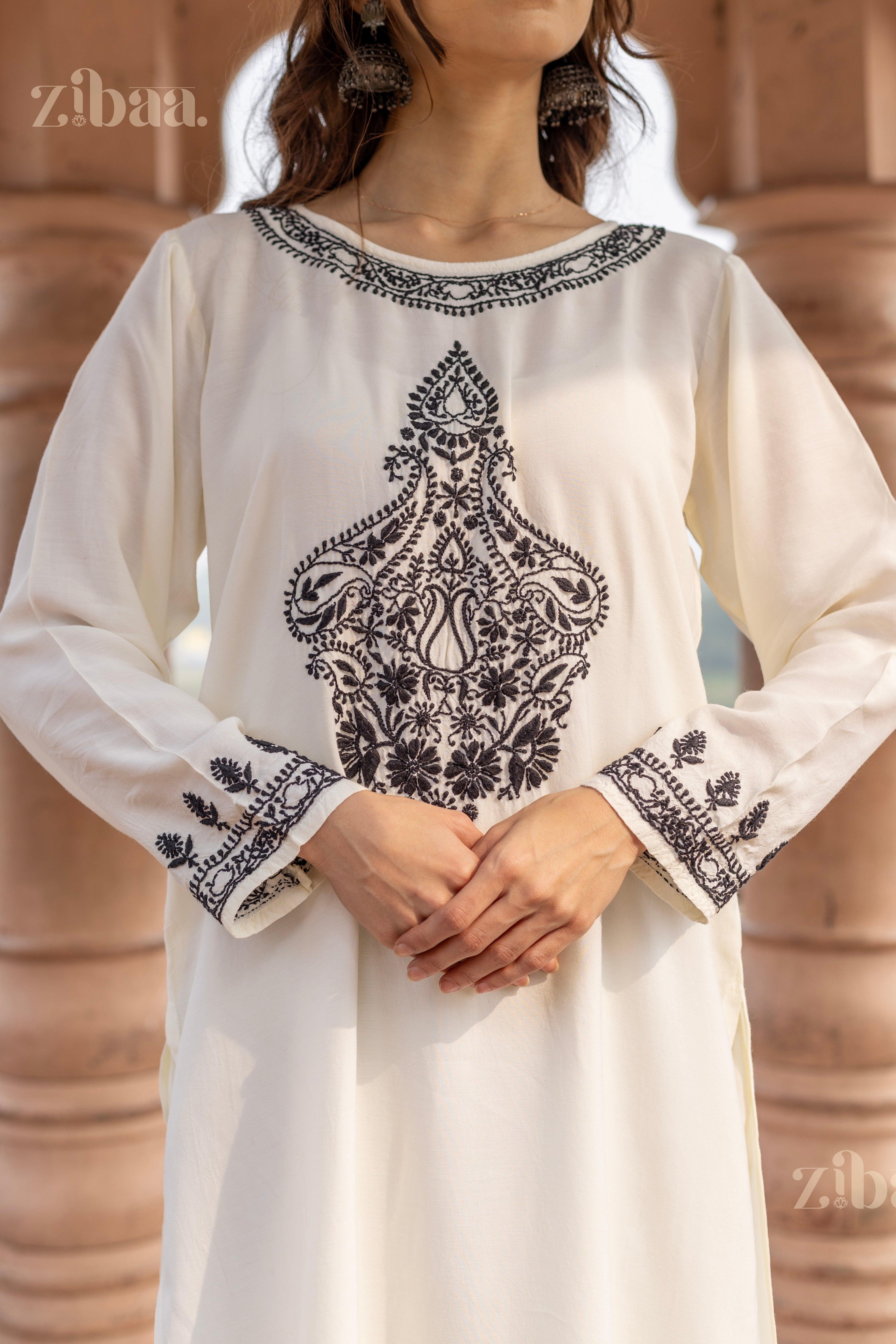 Front view of a woman wearing a white Chikankari kurta set with intricate black embroidery, standing outdoors with hands gently clasped.