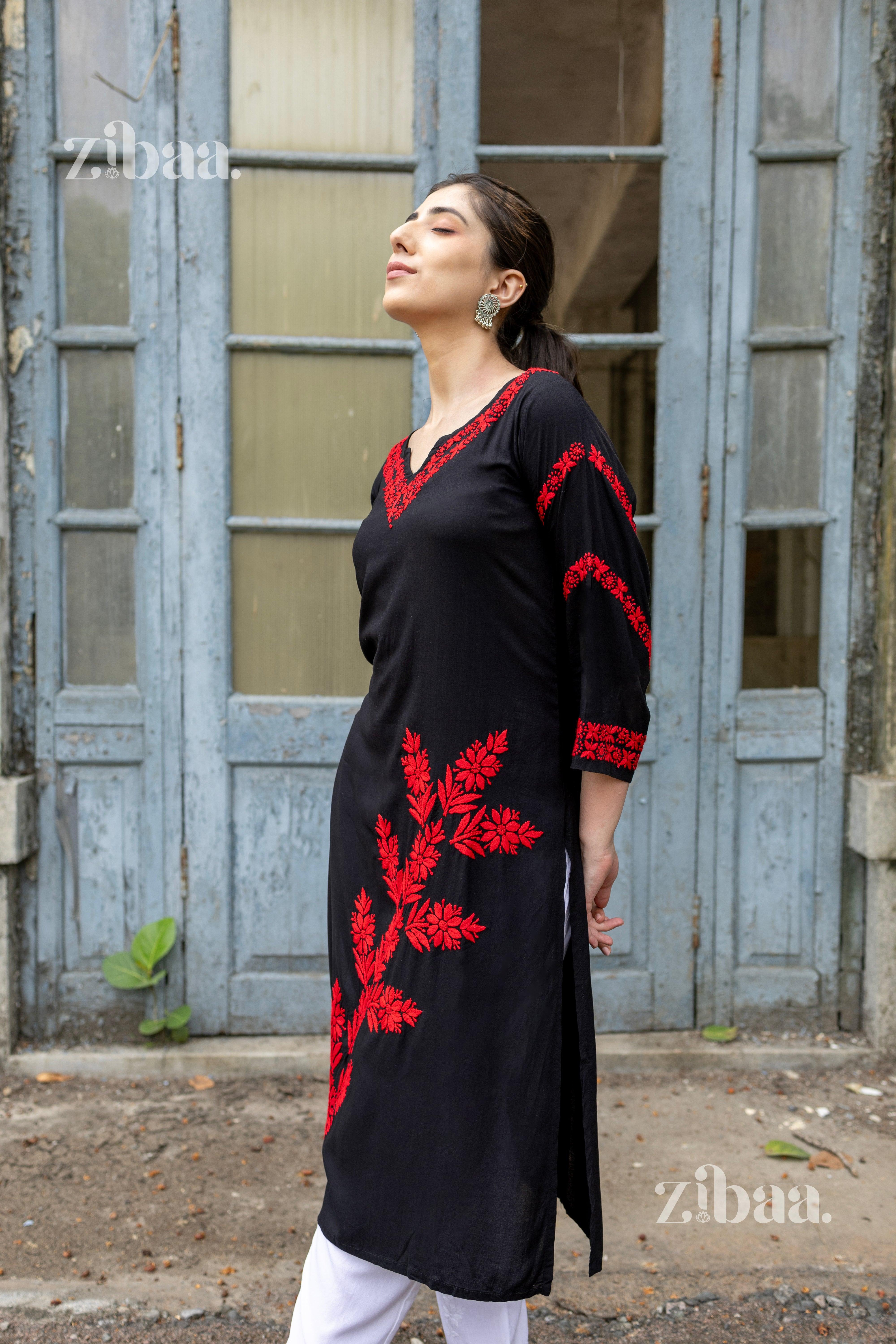 A woman wearing a black kurti with red floral embroidery stands with her hands behind her back, in front of a rustic blue door.