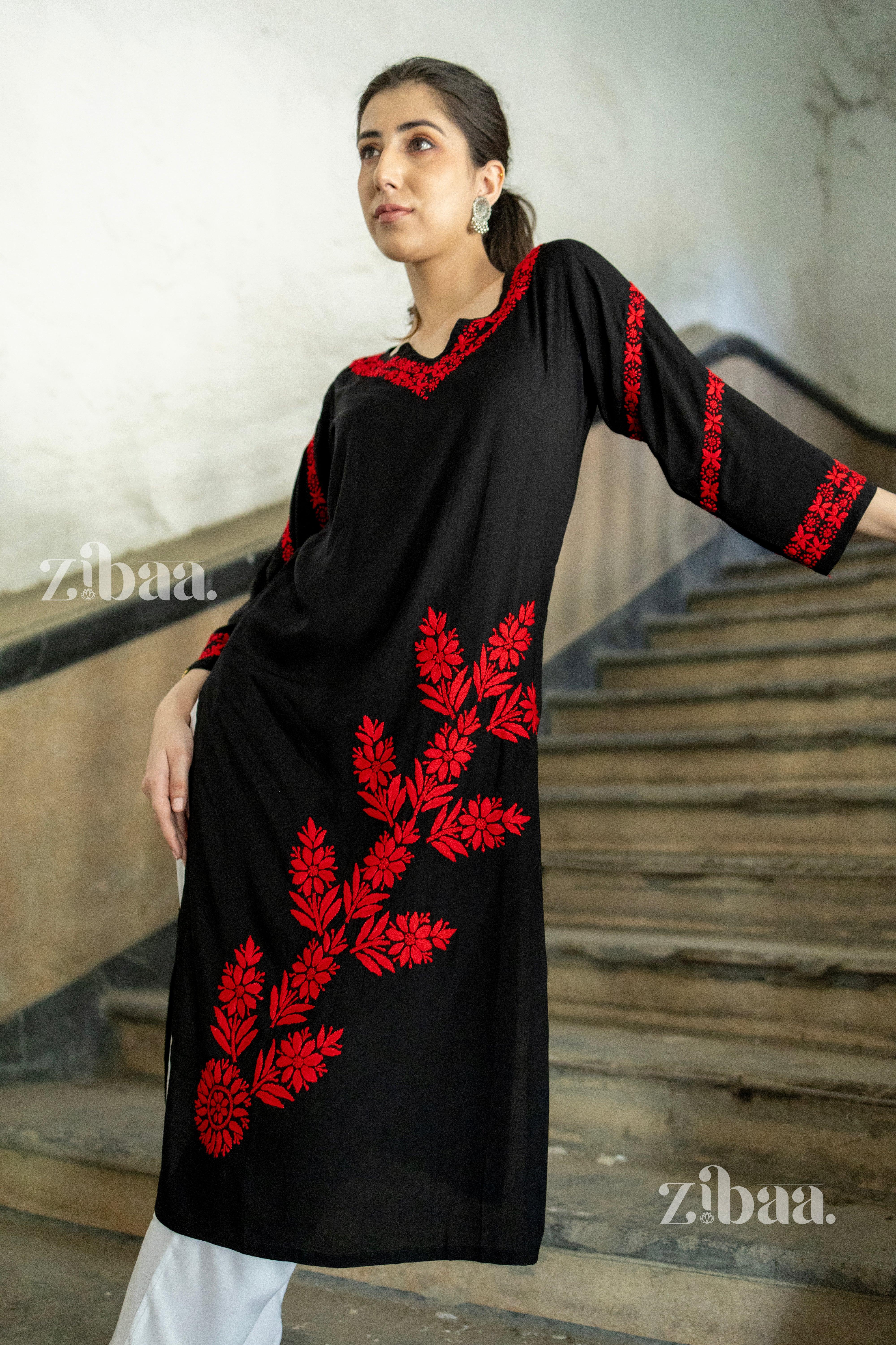 A woman wearing a black kurti with red floral embroidery stands on a staircase, leaning slightly on the railing, looking to the side.