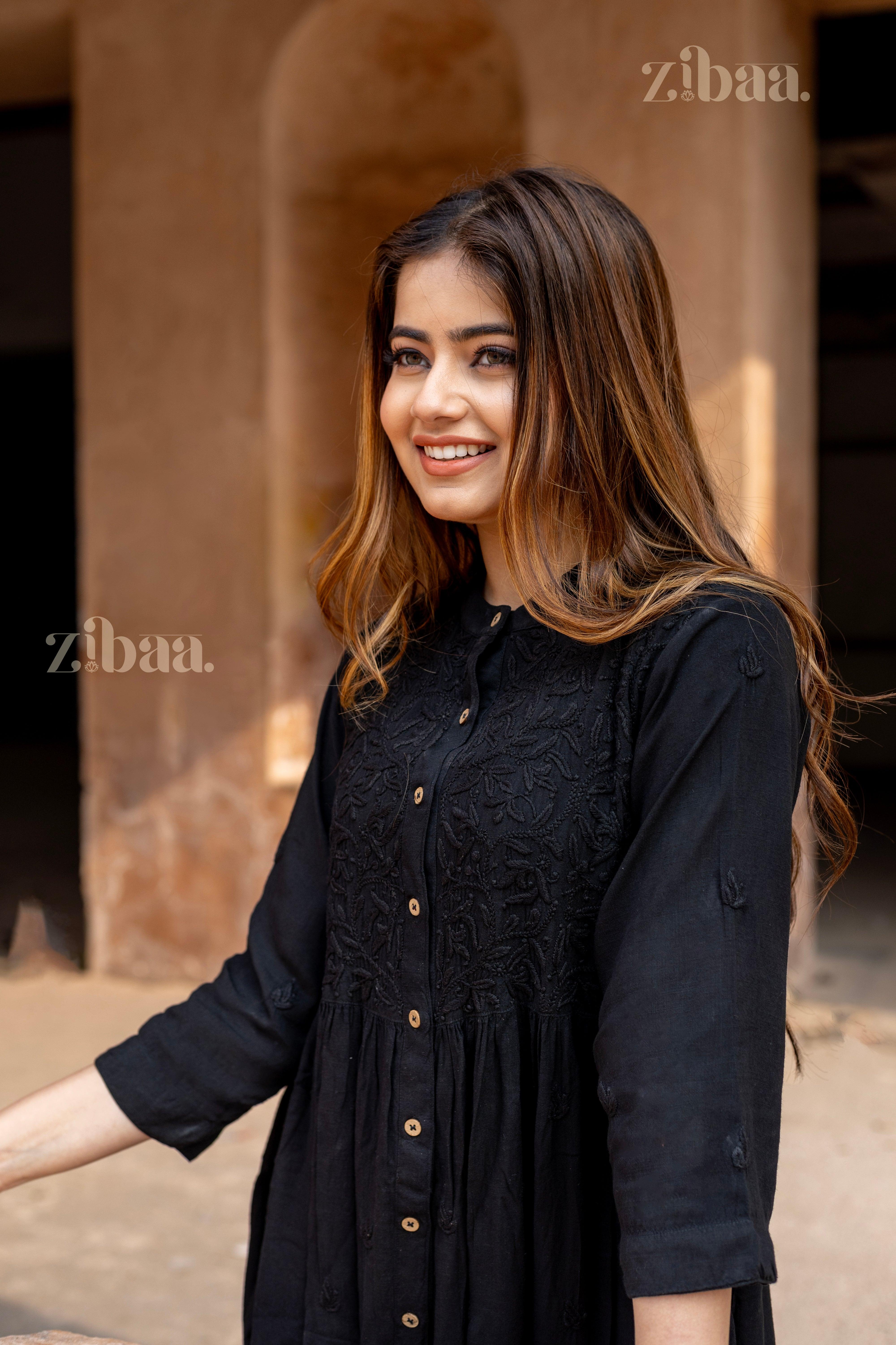 A woman smiles while standing in a traditional setting, wearing a black Chikankari Kurti with embroidery and button-down details.