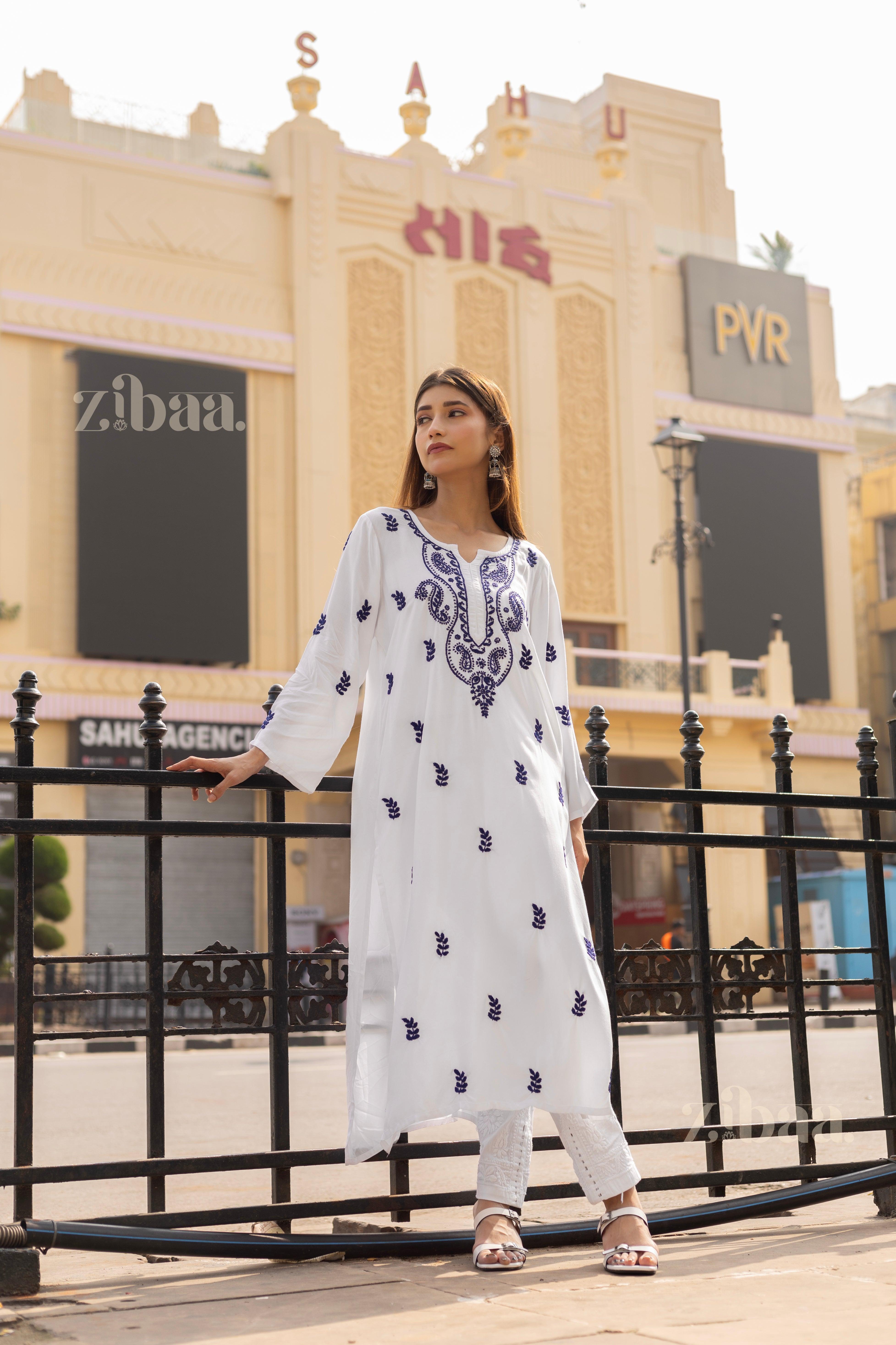 A woman styles a white Chikankari kurti with blue embroidery, paired with white leggings, posing outdoors near a heritage building