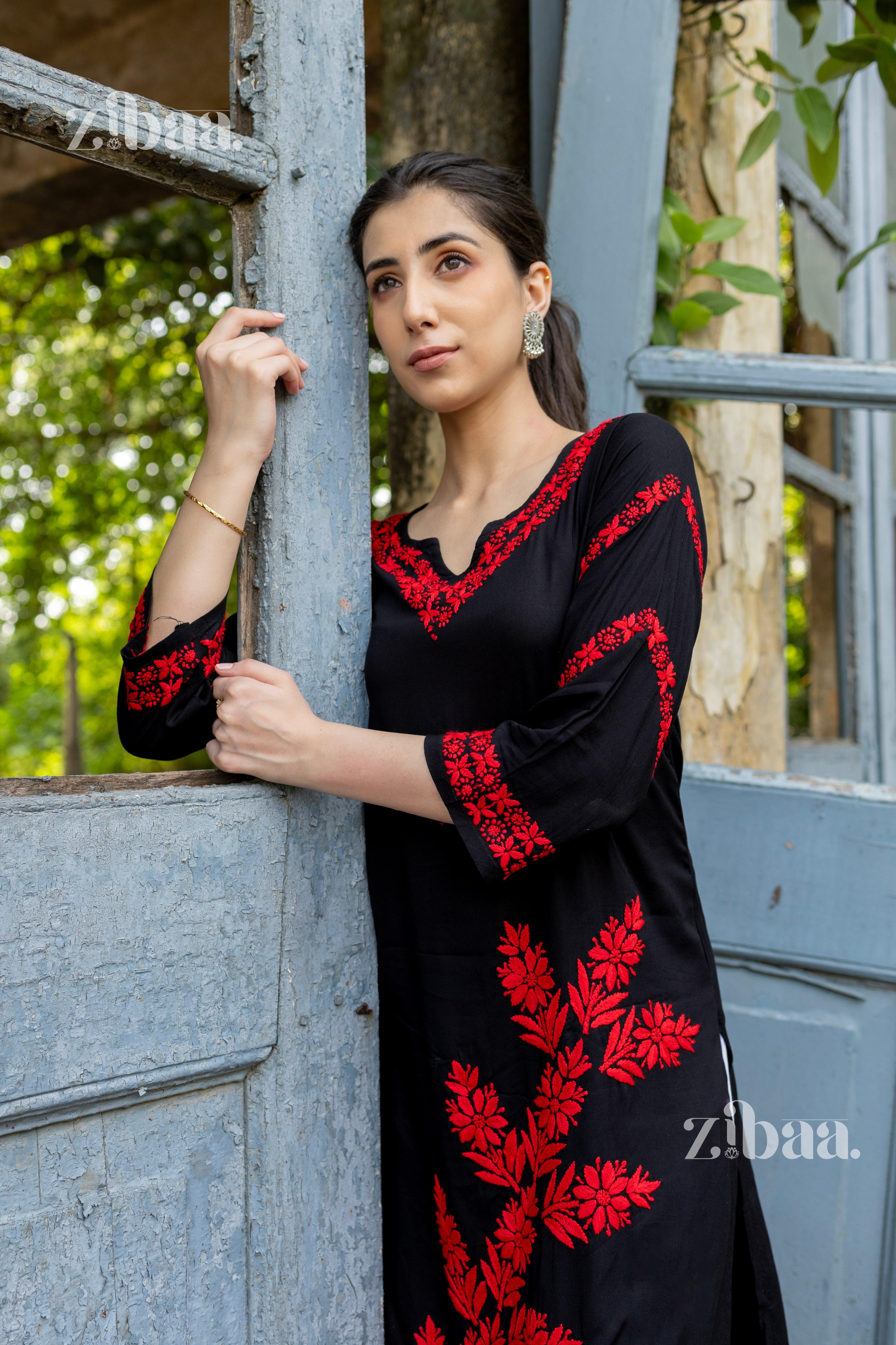 A woman wearing a black Chikankari kurti with red embroidery stands leaning against a rustic blue door, looking away thoughtfully.