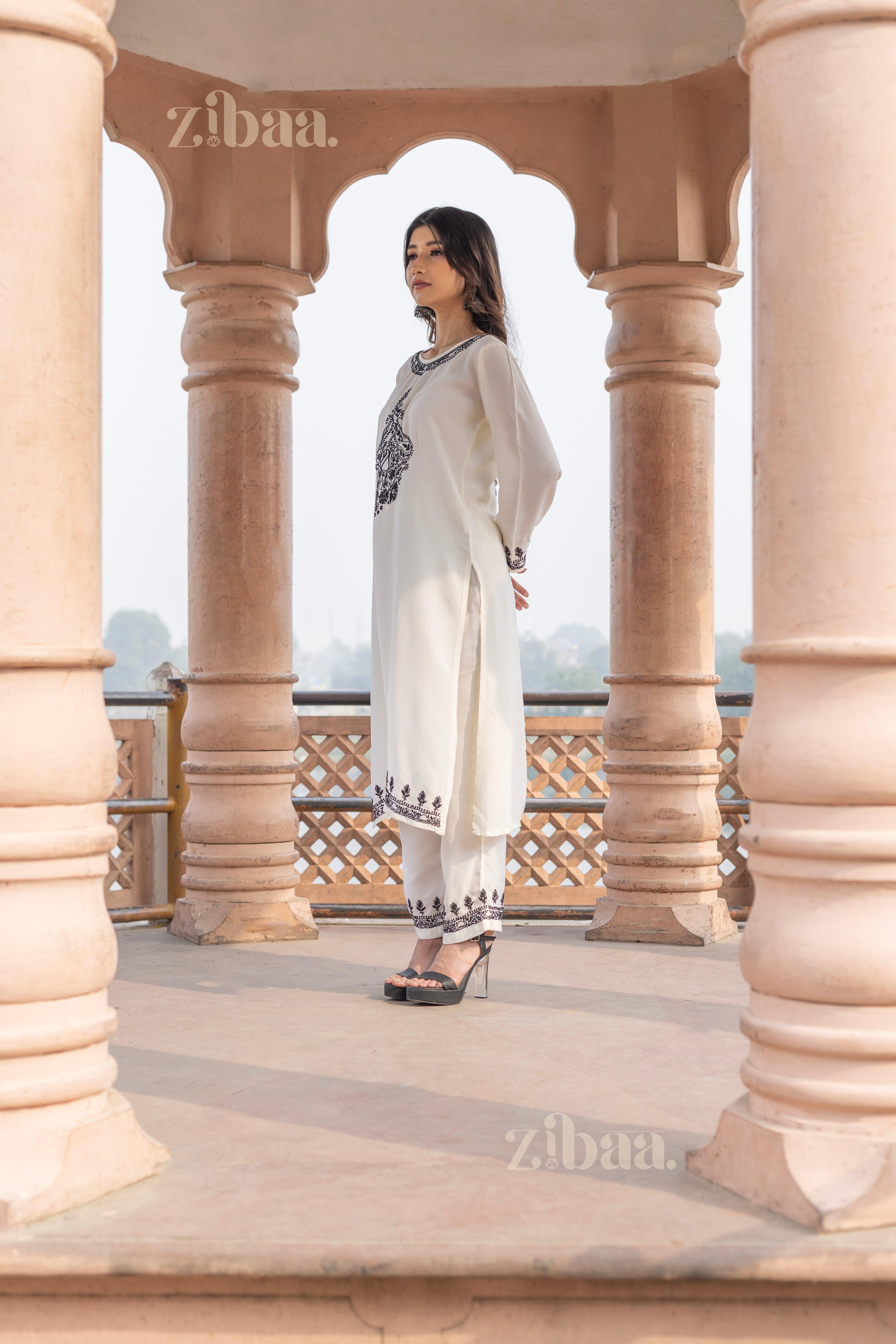 A woman wearing a white Chikankari kurti with black embroidery stands between ornate pillars, gazing into the distance outdoors.