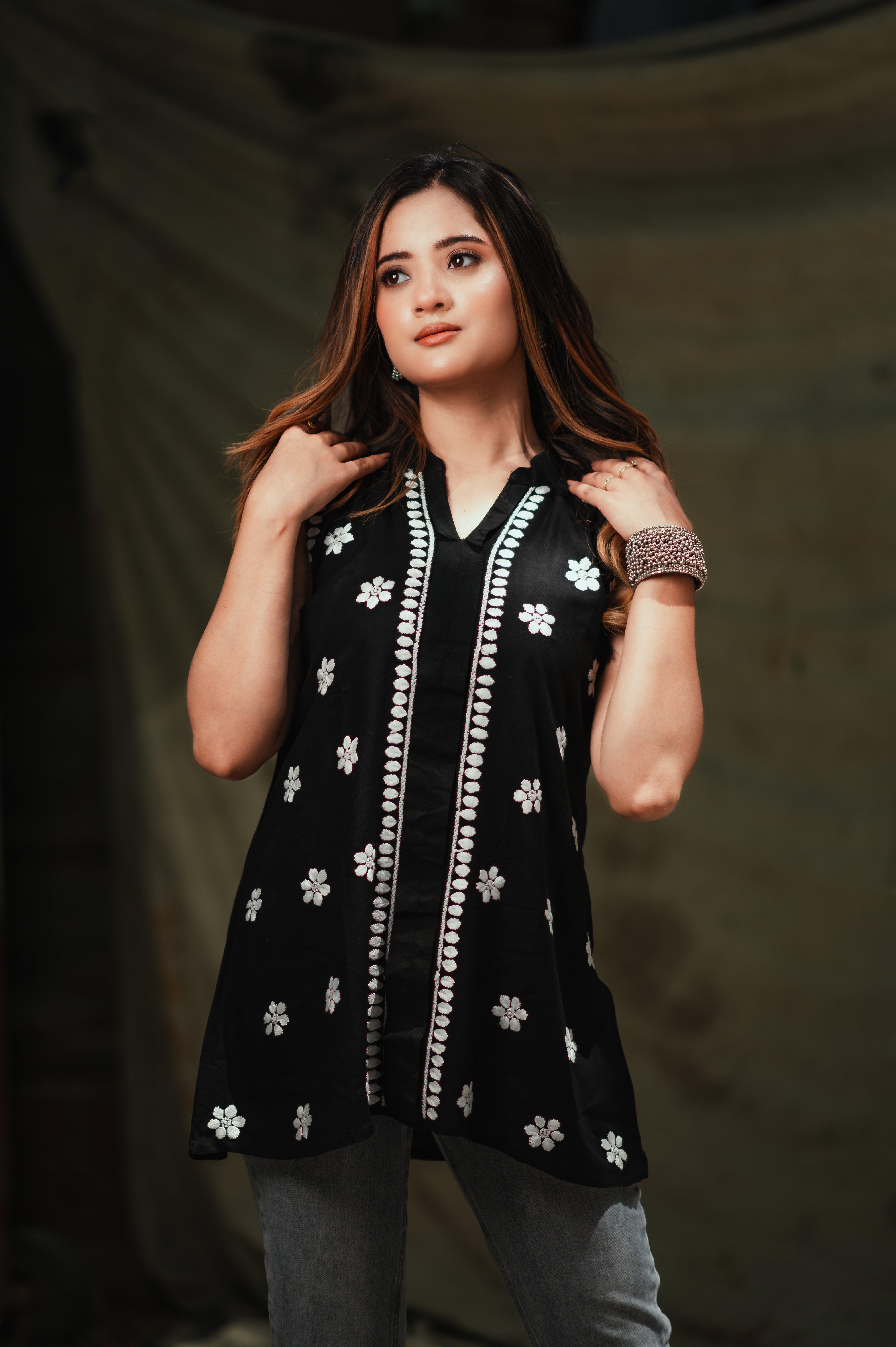 A woman wearing a black Chikankari top with white floral embroidery stands confidently, holding her hair, looking to the side.