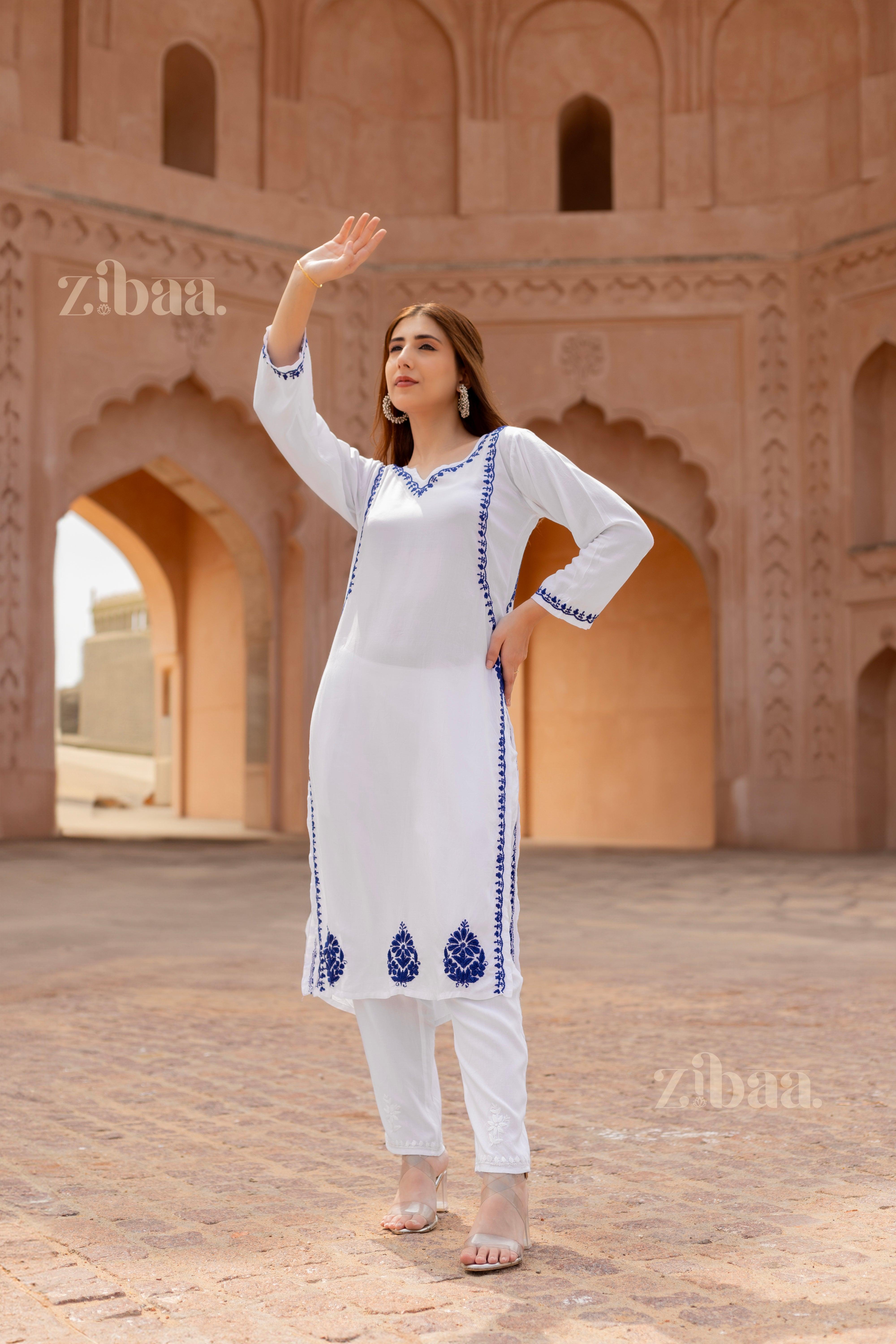 Woman posing confidently with one arm raised, wearing a white Chikan Kurti with blue embroidery, standing in a heritage-inspired setting