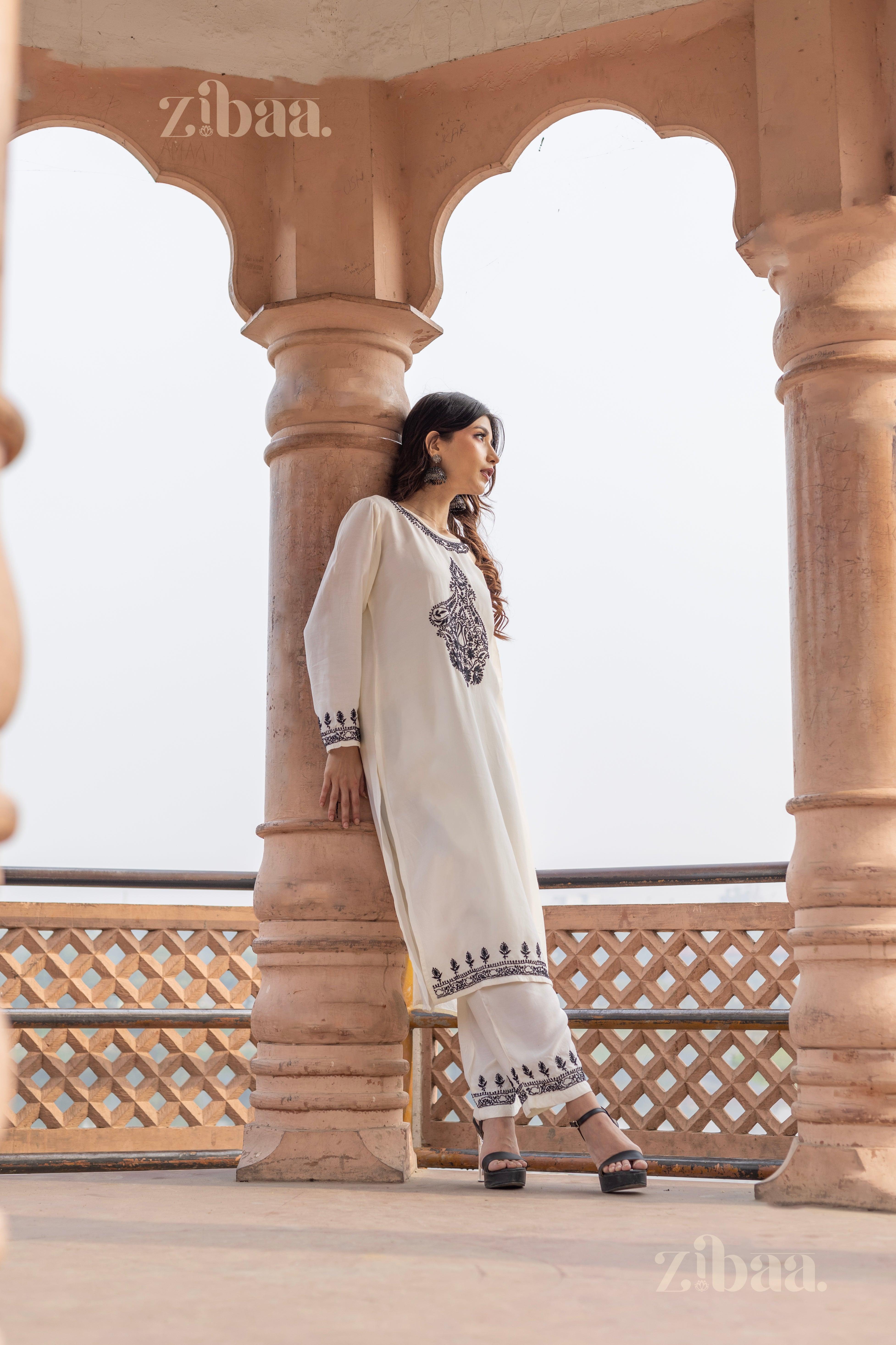 A woman wearing a white Chikankari kurta set with black embroidery leans against a pillar, gazing to the side, in a heritage pavilion.