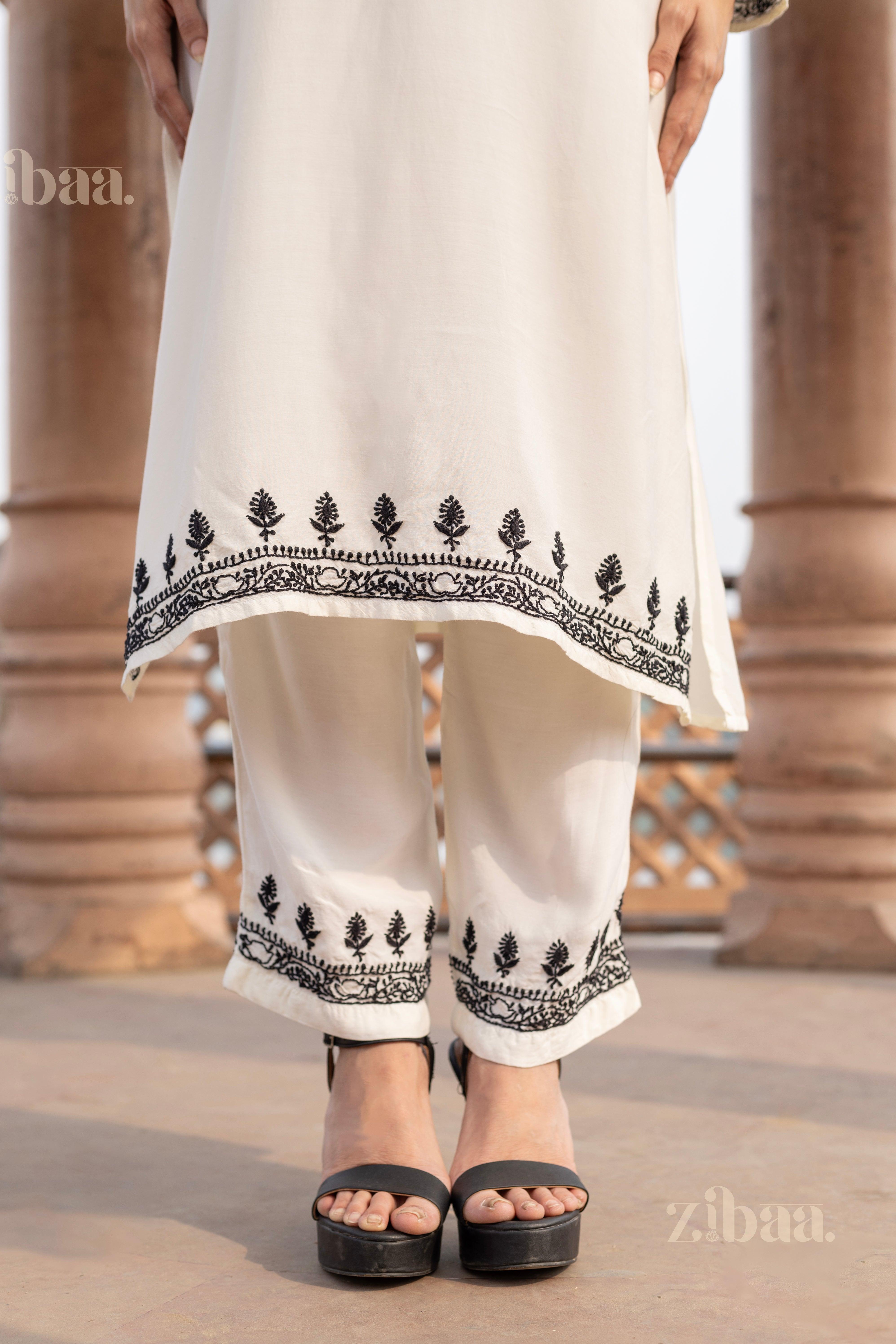 Close-up of a white kurta set for women featuring intricate black embroidery on the hem and pants, as the woman stands in black heels.