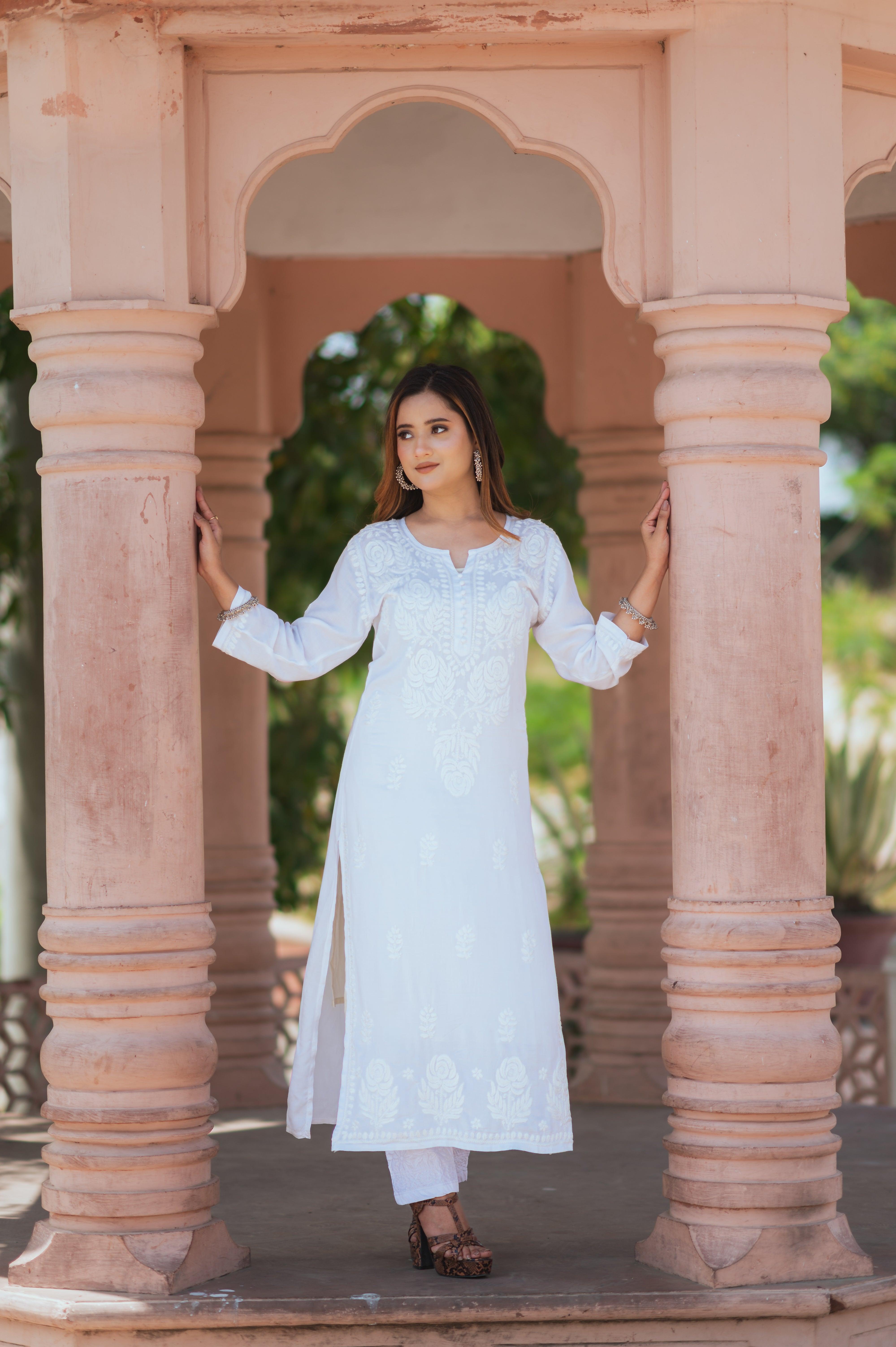 A woman in a white Chikankari kurti with intricate embroidery stands between two pillars, holding with her hands, looking to the side.