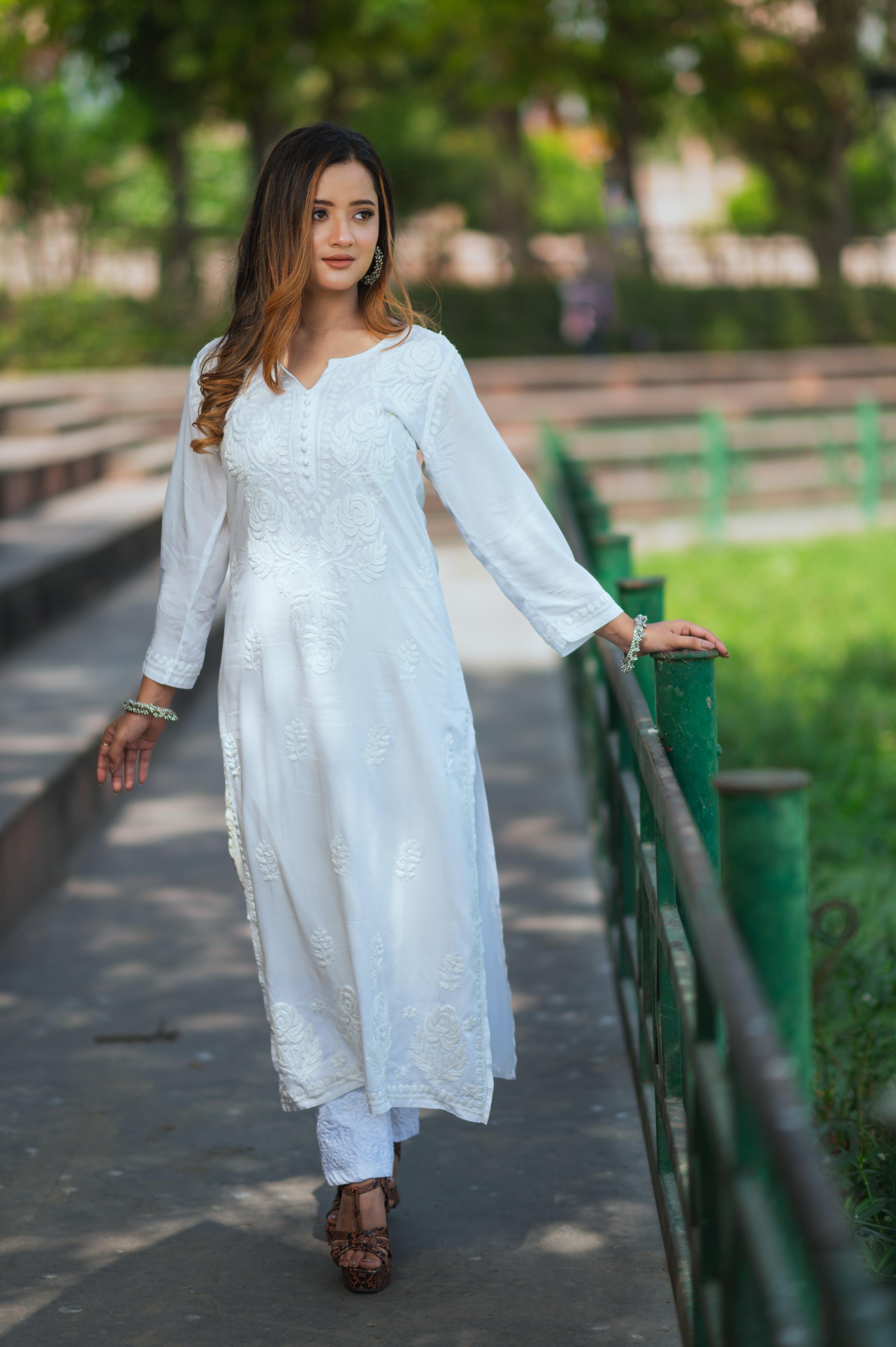 A woman in a white Chikankari kurti with intricate embroidery walks gracefully along a railing, lightly touching it with one hand.