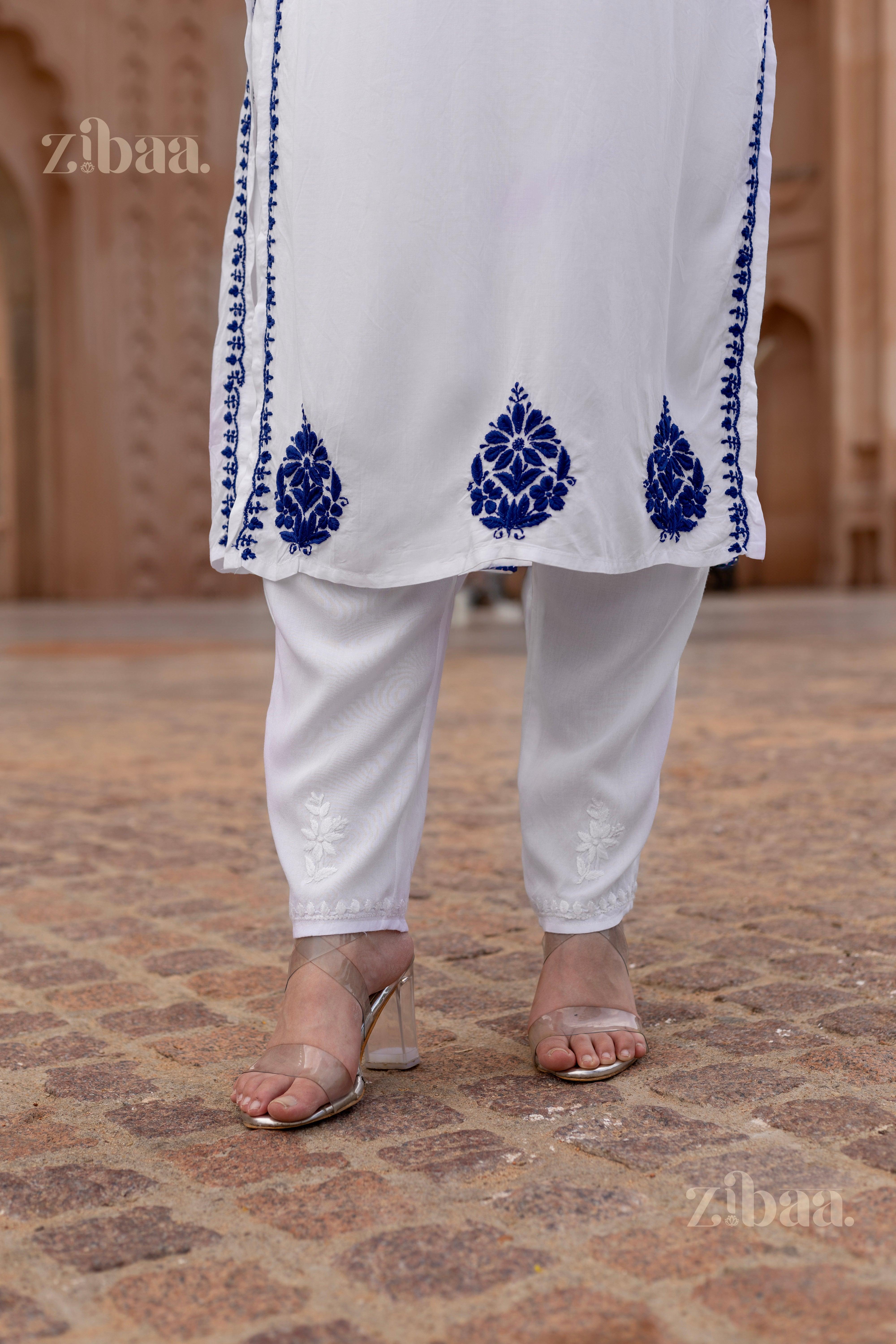 Close-up of a white kurti for women with intricate blue embroidery near the hem, paired with white pants and transparent heels, outdoors