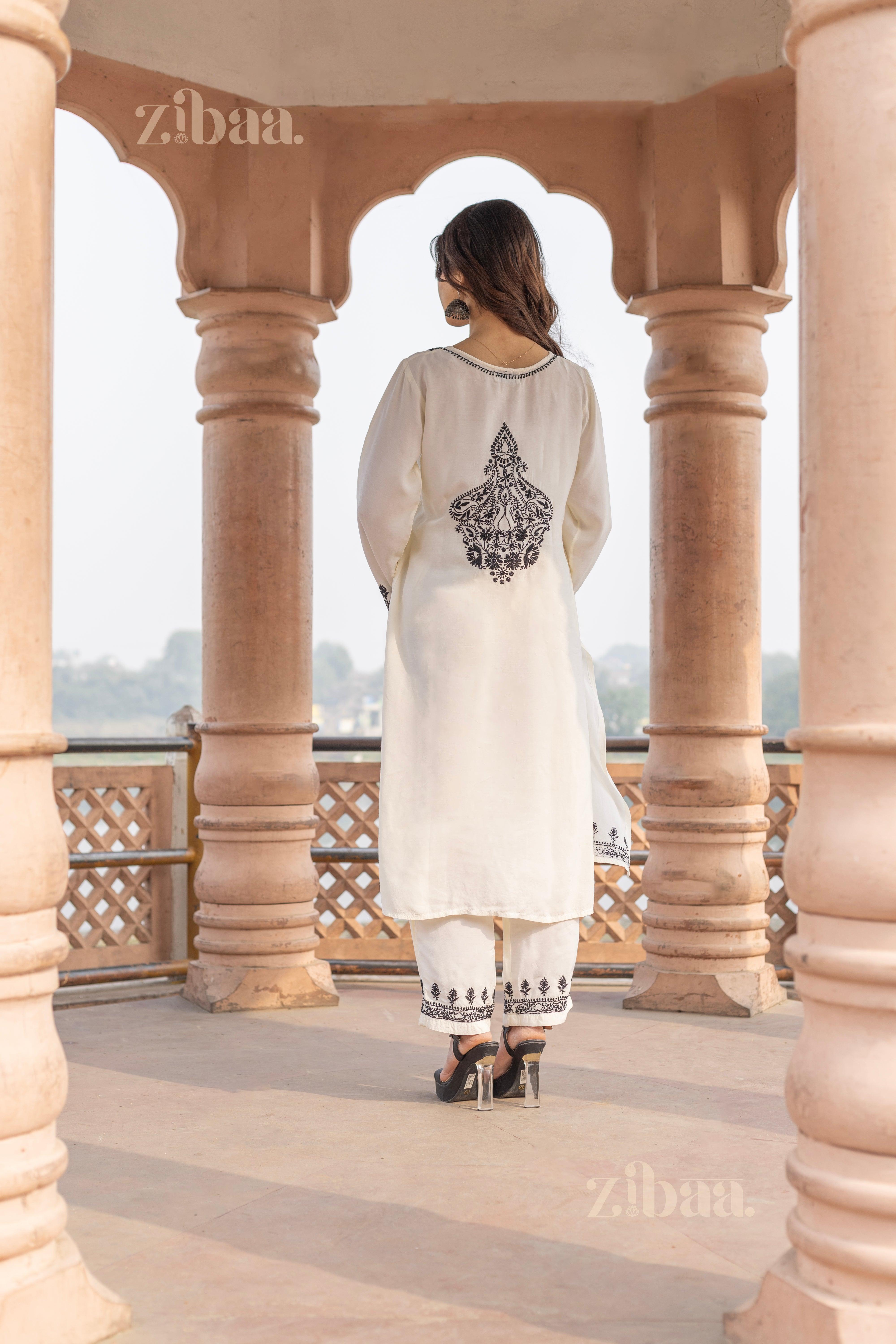 A woman wearing a white Chikankari kurti with intricate black back embroidery stands outdoors between pillars, facing away gracefully.