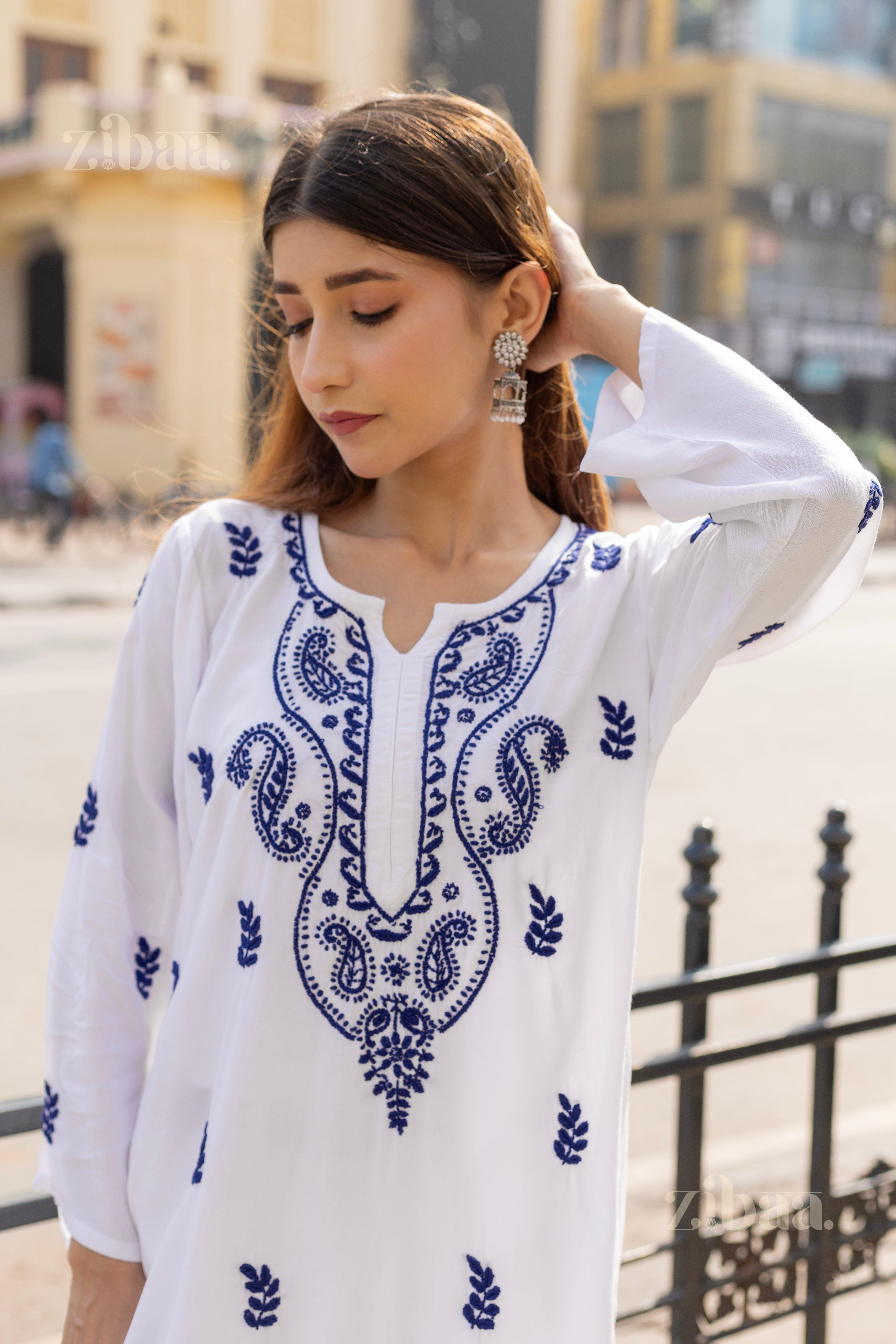 A woman is posing in a white Chikankari kurti featuring intricate blue embroidery, paired with silver earrings in an outdoor area.