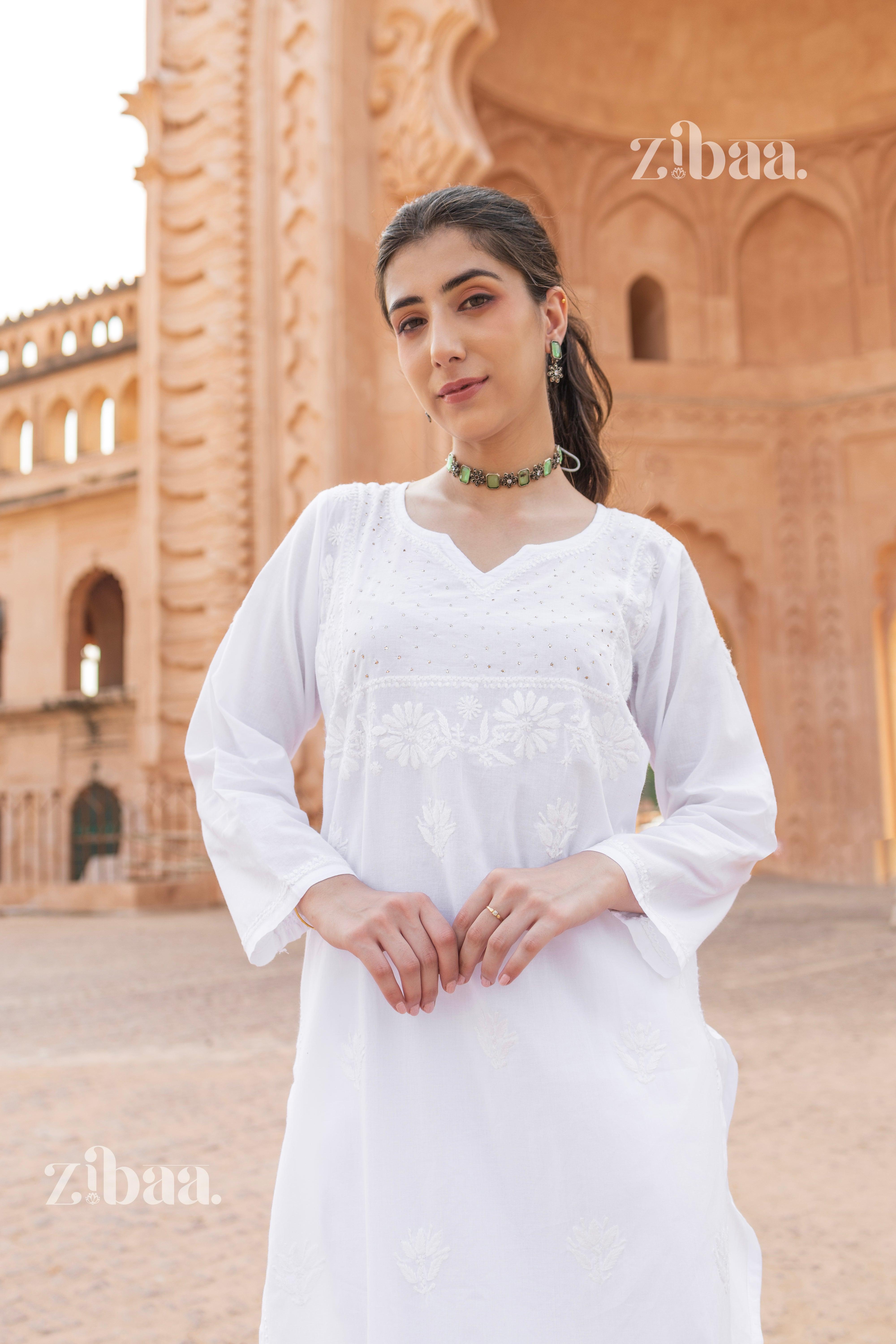 A woman in a white chikan kurti adorned with floral embroidery, paired with a green choker necklace, posing in a heritage setting.