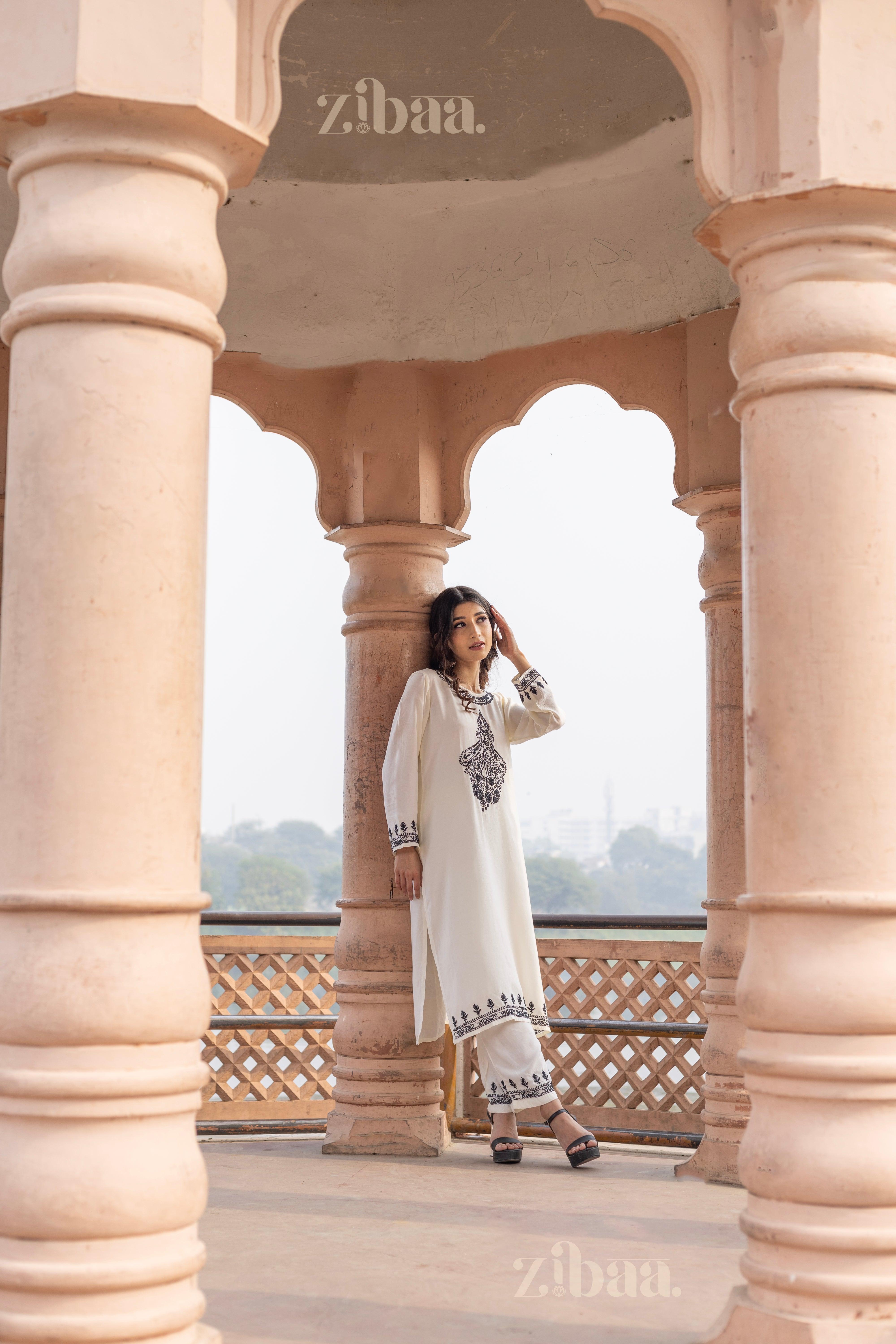 A woman wearing a white Chikankari kurta set with black embroidery stands outdoors between pillars, adjusting her hair and posing.