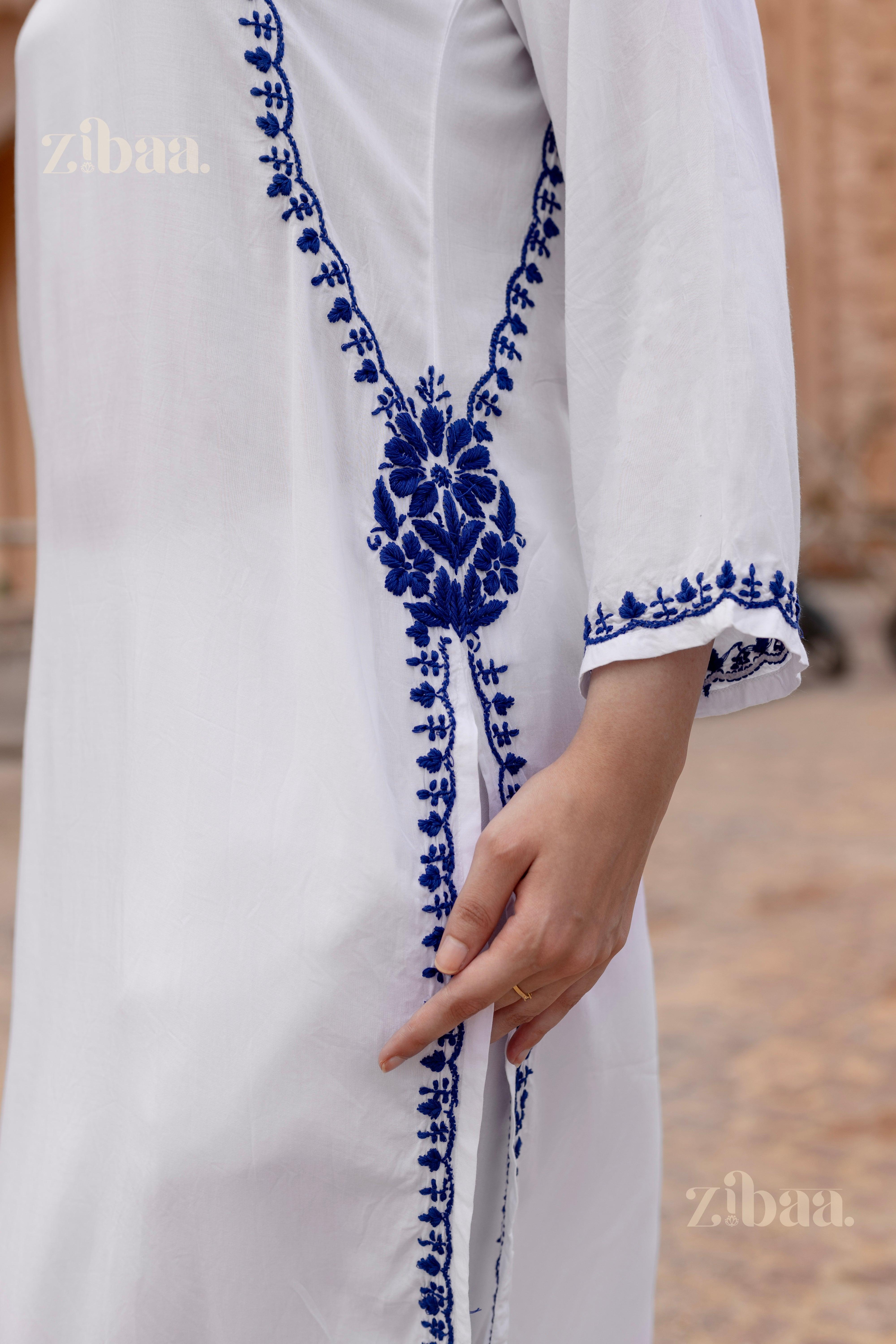 Model walking outdoors wearing a white kurti for women with intricate blue embroidery, posing in front of a heritage backdrop