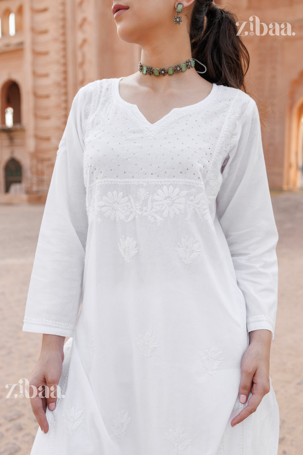 A girl in a white kurti for women with delicate embroidery, paired with white pants and heels, posing in a heritage-inspired backdrop.