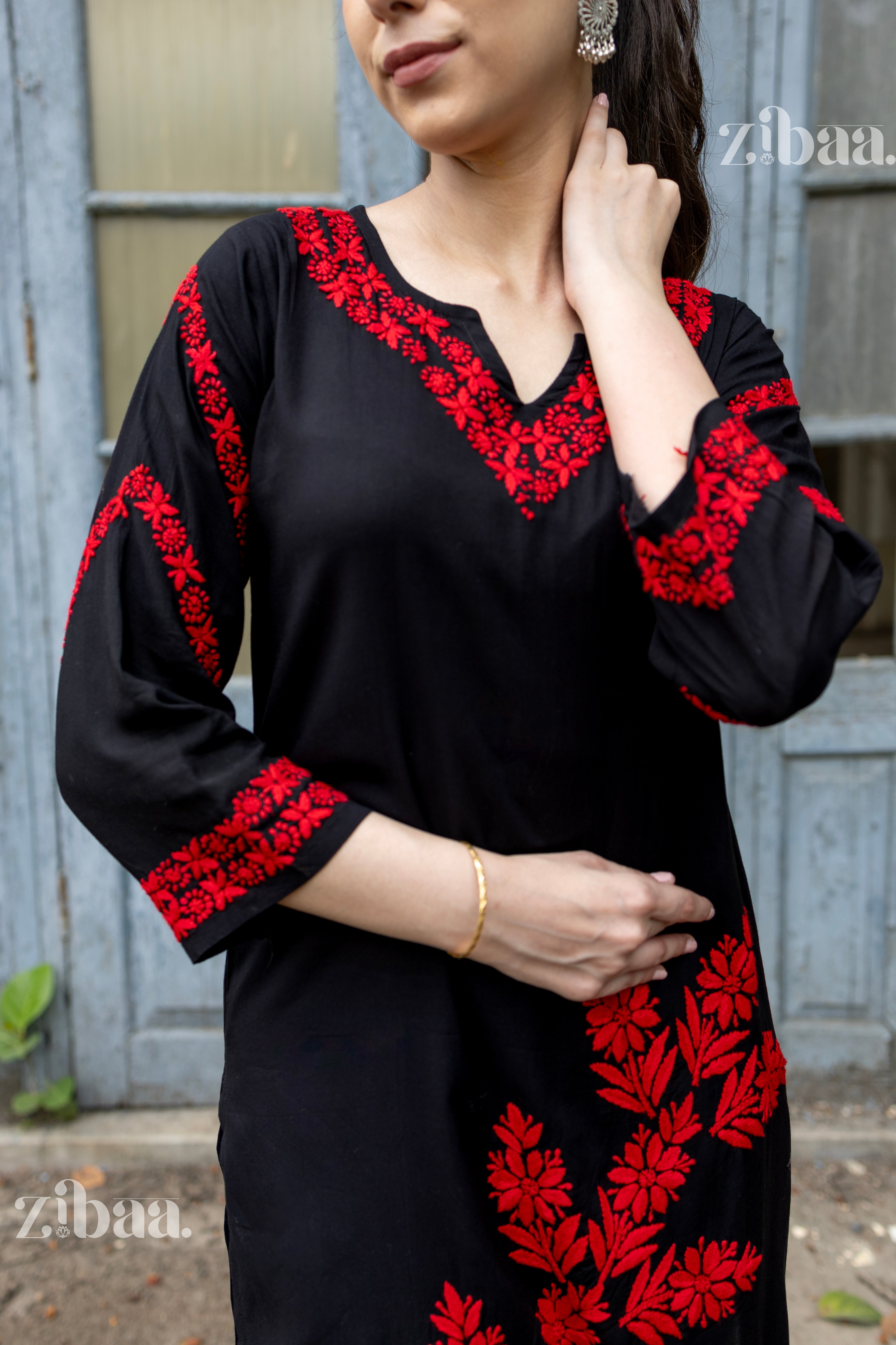 A woman in a black kurti for women featuring vibrant red floral Chikankari embroidery, posing elegantly by a rustic door, styled.
